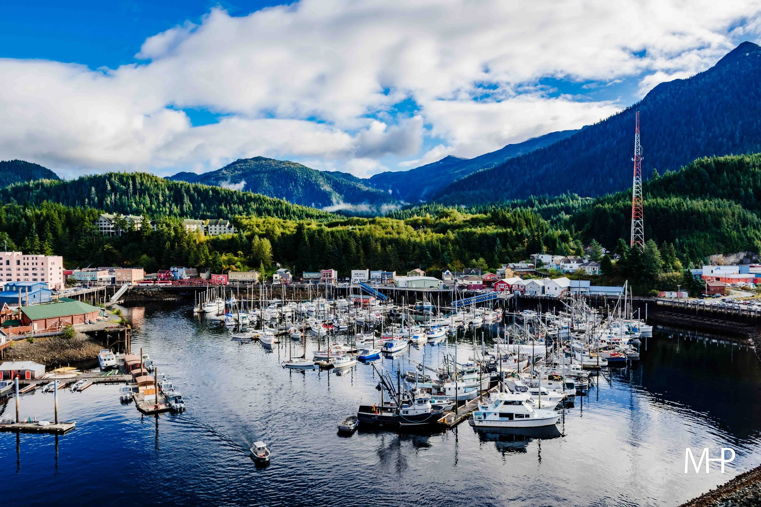 A marina with numerous sailboats and yachts docked, surrounded by colorful buildings along the waterfront, set against lush green forested hills and mountains under a partly cloudy sky.