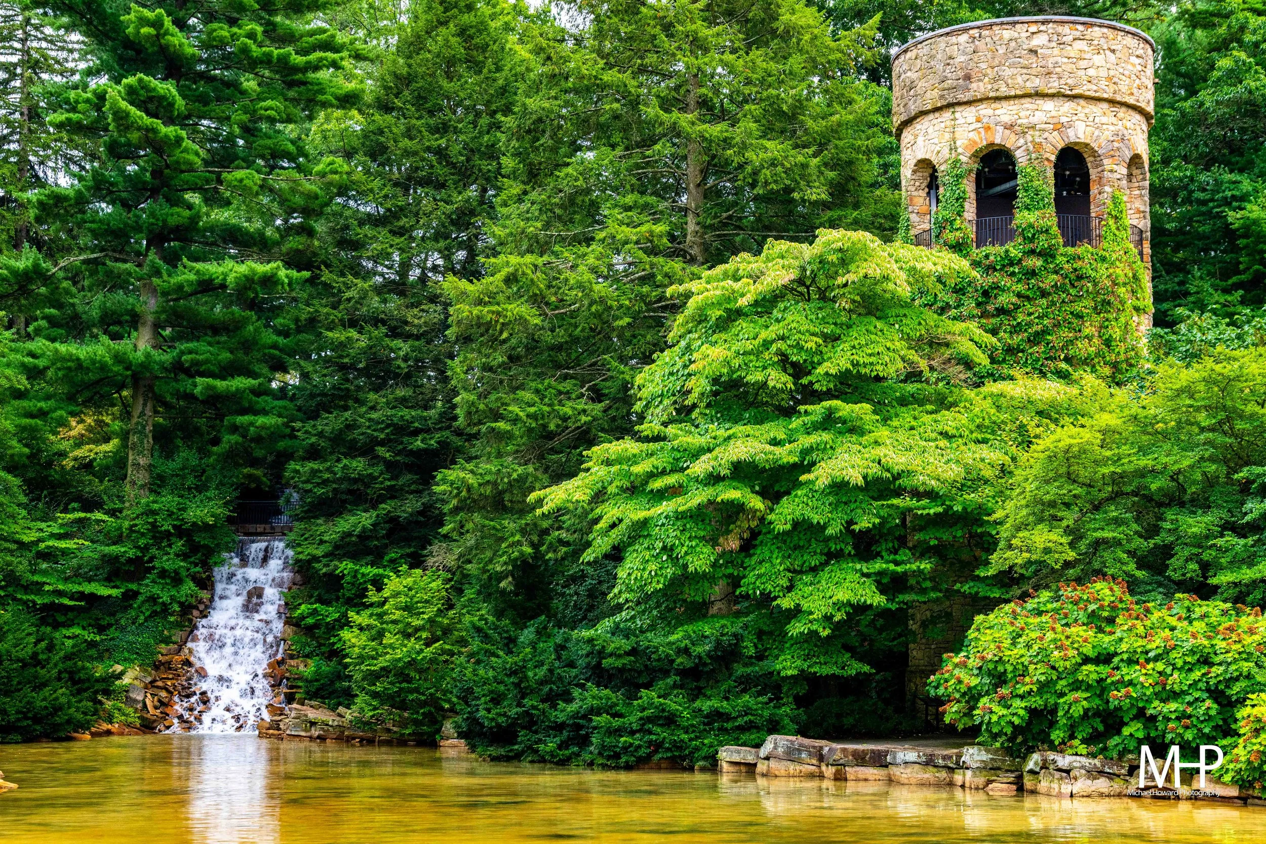 Longwood Gardens, A lush, green forest with a small waterfall cascading into a river, featuring a stone tower with arched openings covered in vines.