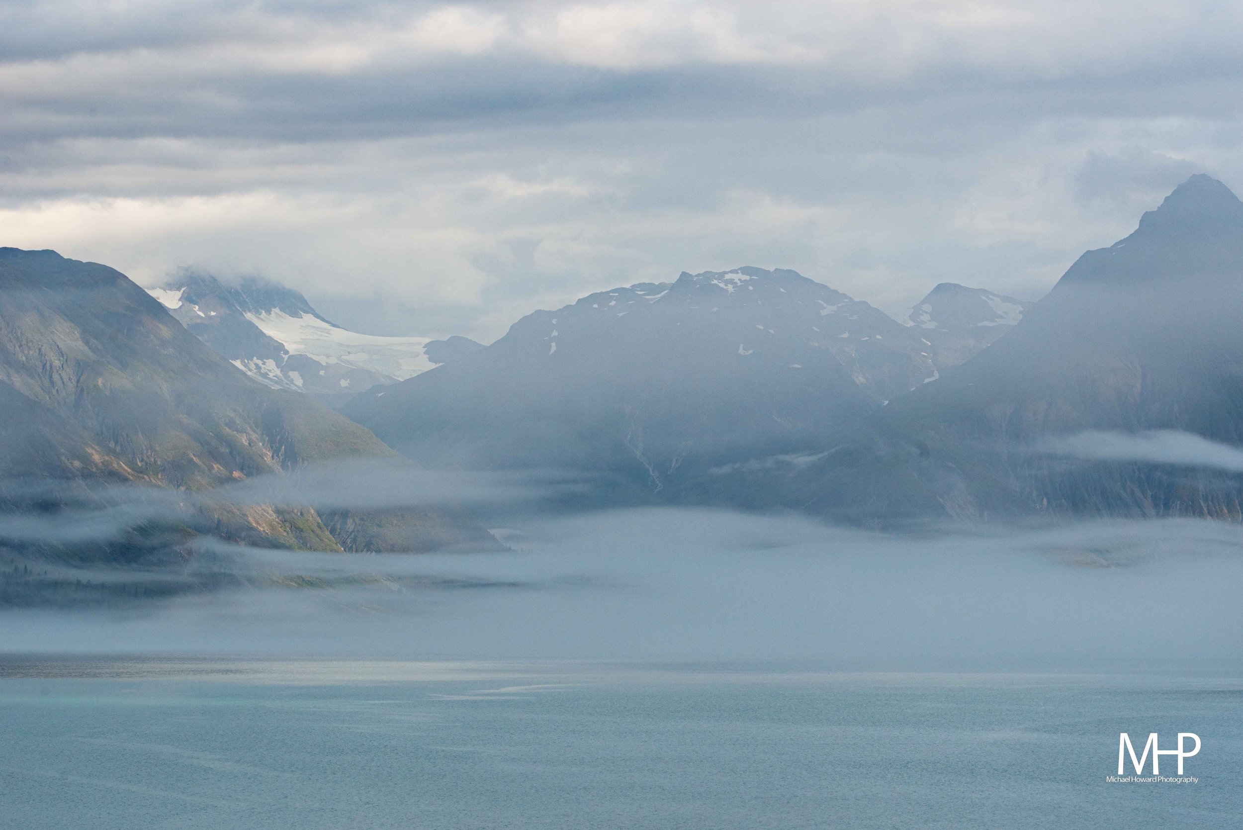 Mountains with snow patches, covered partially by mist and clouds over a body of water.