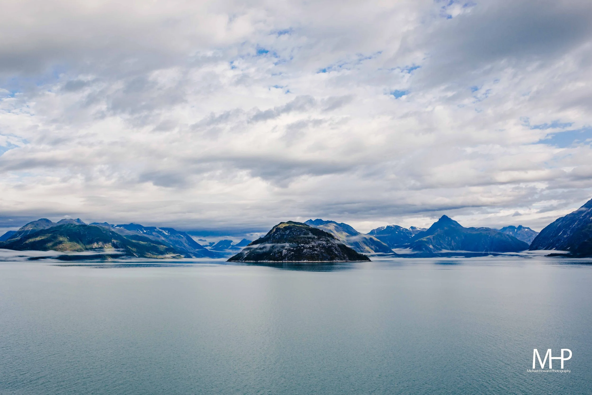 Scenic view of a fjord with calm water, surrounded by snow-capped mountains under a cloudy sky.