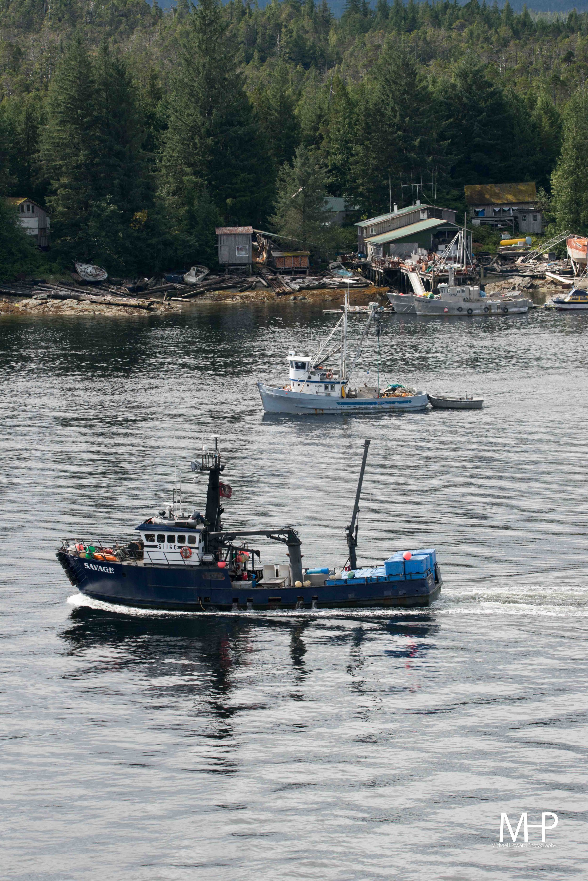 A coastal scene with a large boat named 'SAVAGE' moving through the water in the foreground. Several other boats are anchored nearby, and there are houses and dense trees on the shoreline in the background.