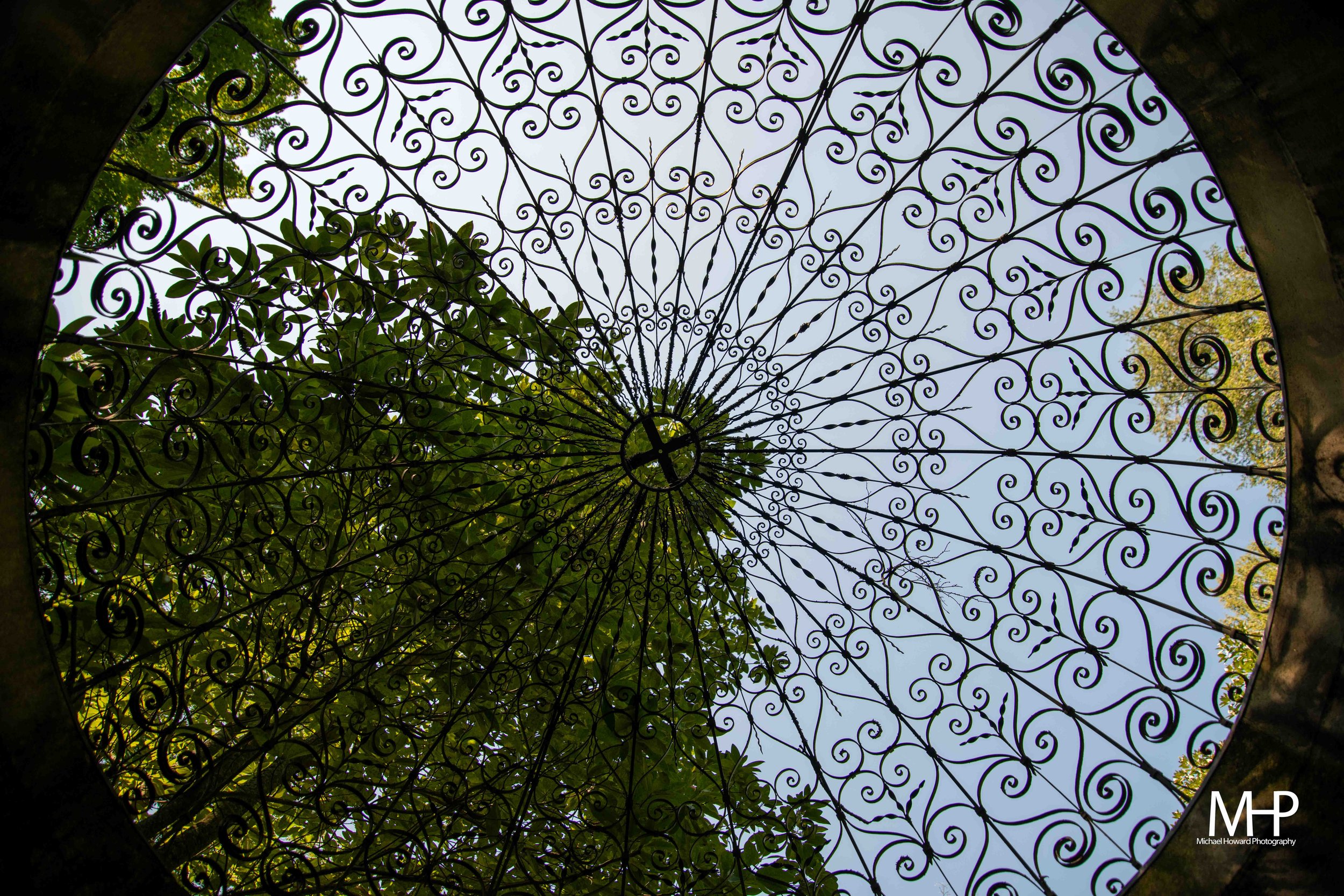 Looking up through an ornate wrought iron overhang with swirling patterns, with blue sky and green trees visible in the background.