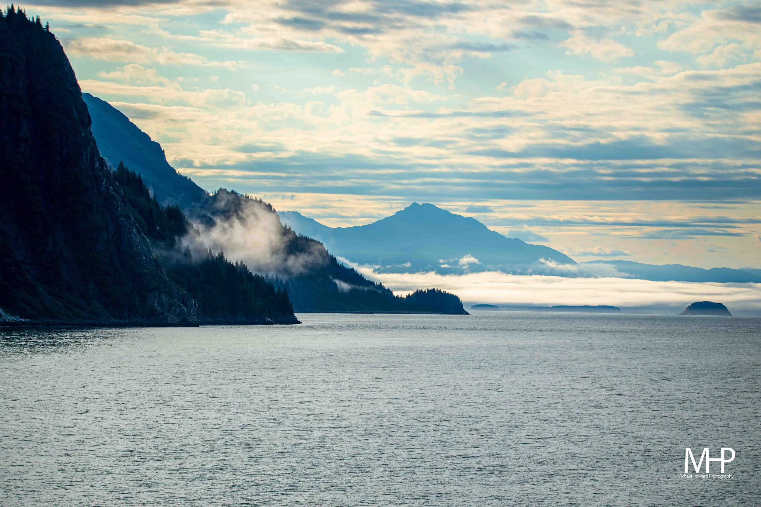 Calm body of water with forested mountain cliffs on the left side, mist rising from the trees, and distant mountains in the background under a partly cloudy sky.