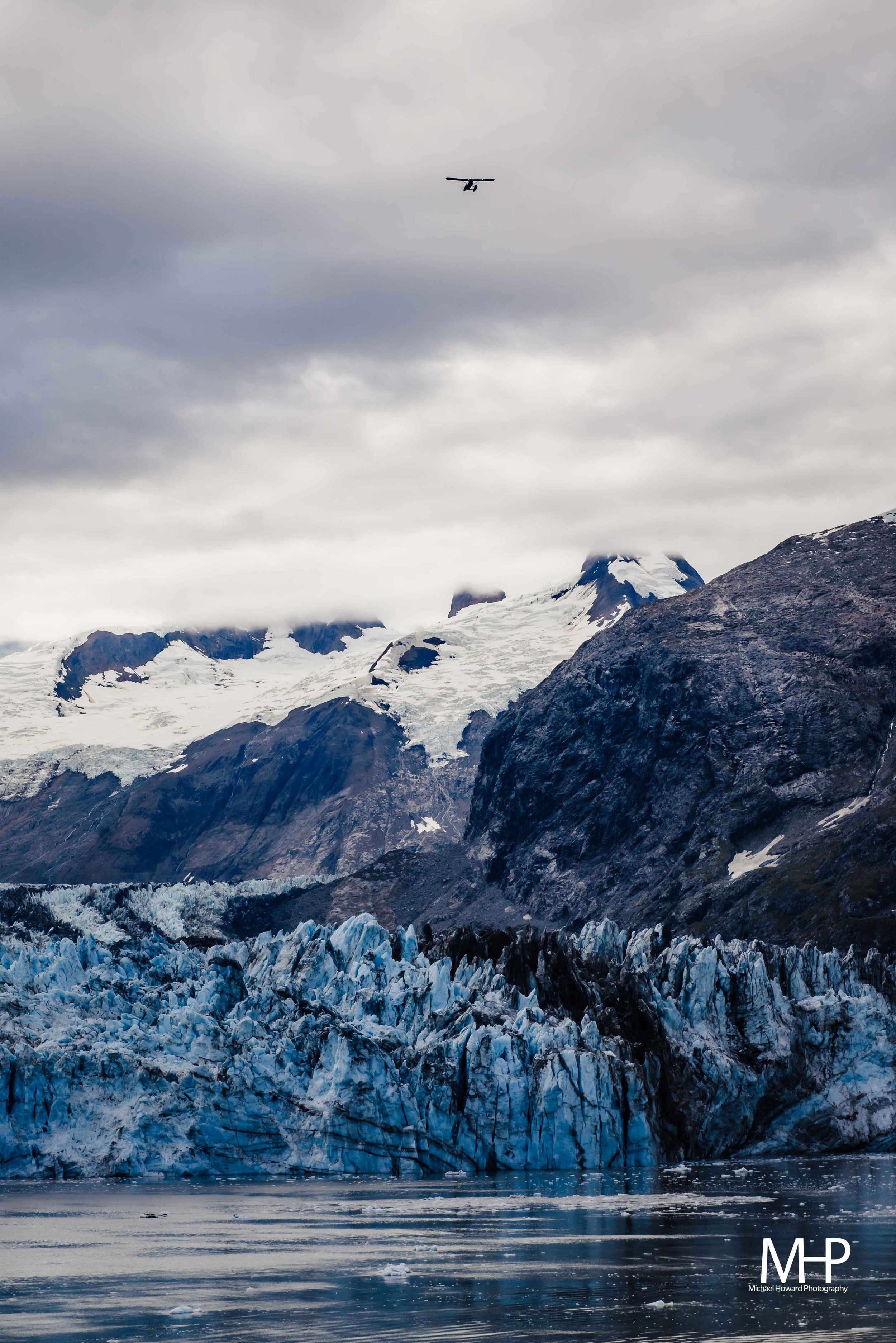 A glacier with icy blue and white crevasses extending into a body of water, surrounding snow-capped mountains and a cloudy sky, with a flying airplane in the distance.