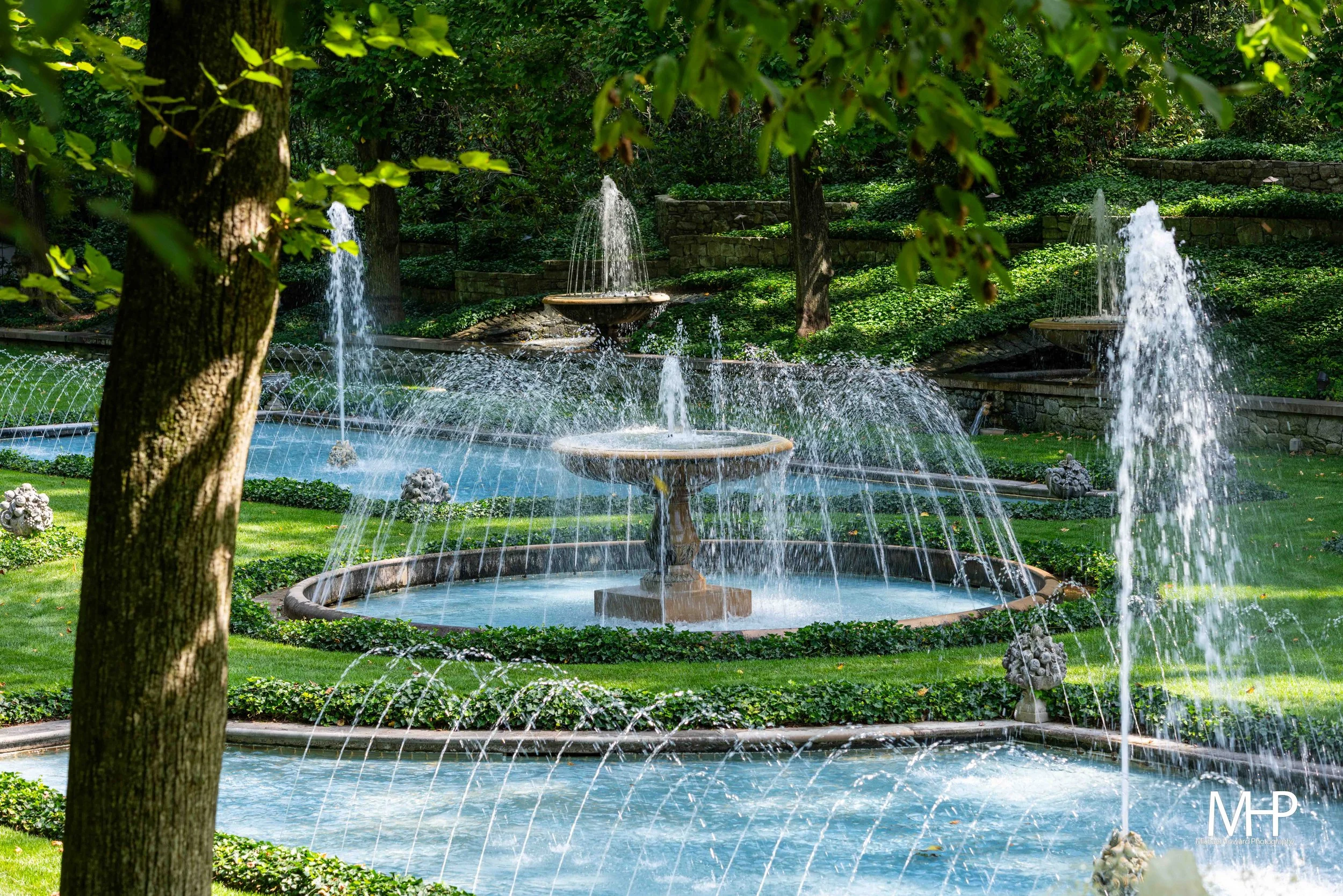 Fountain with multiple streams of water in a garden surrounded by trees and green shrubbery.
