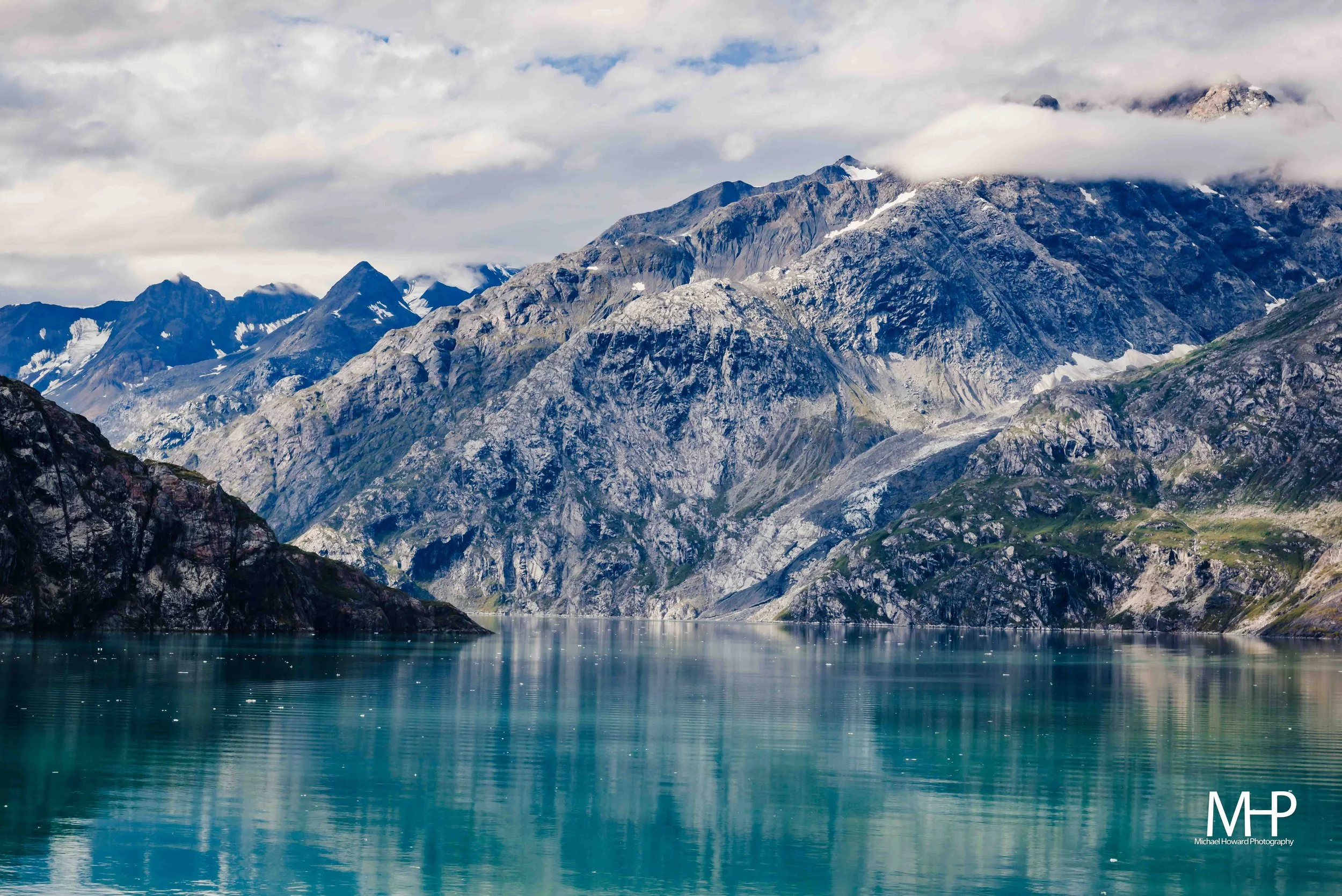 A scenic landscape of a mountain range with snow patches, clouds hovering over the peaks, and a calm blue-green body of water below.