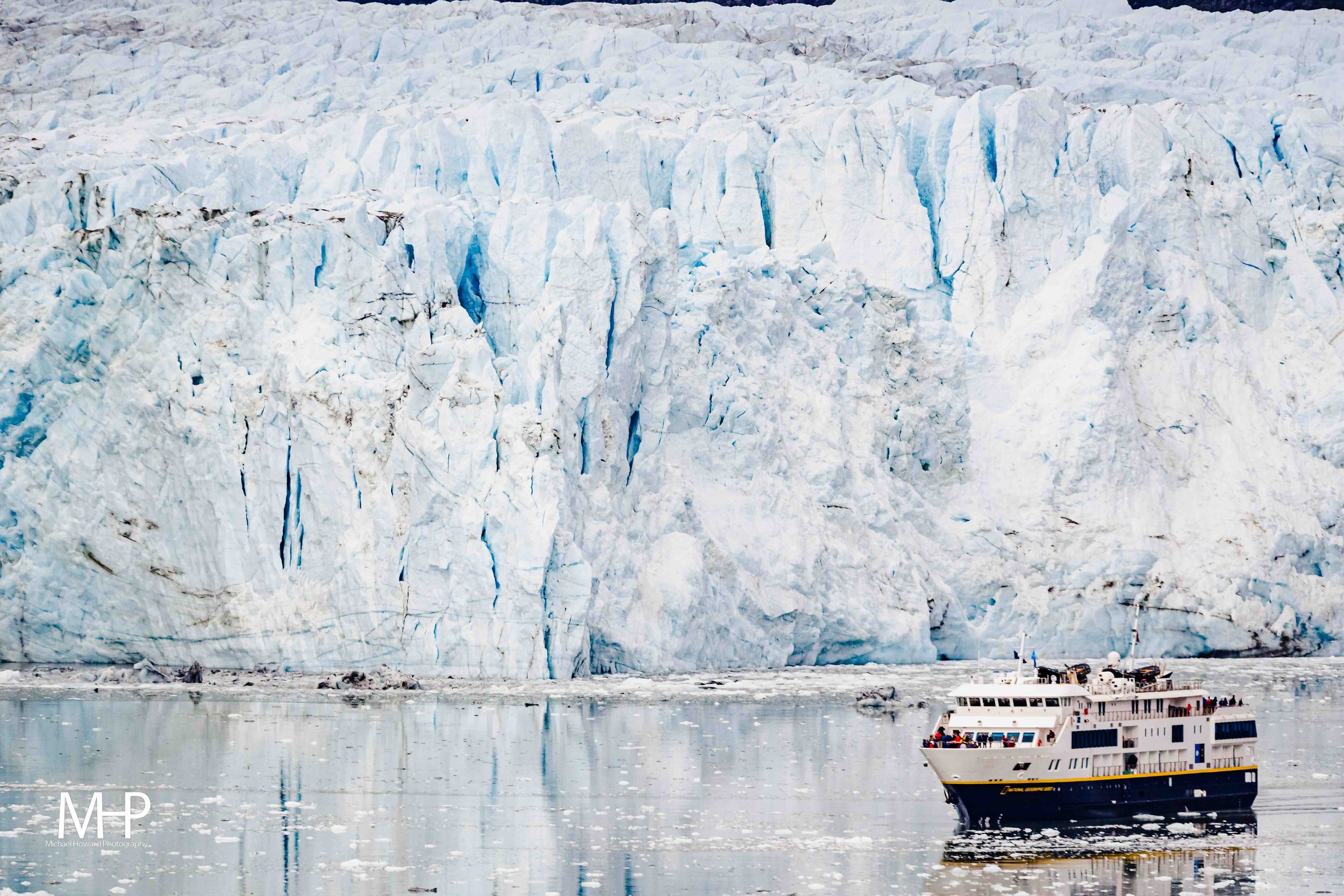 A large glacier with blue ice crevasses, a boat with passengers sailing nearby, and a partly cloudy sky.