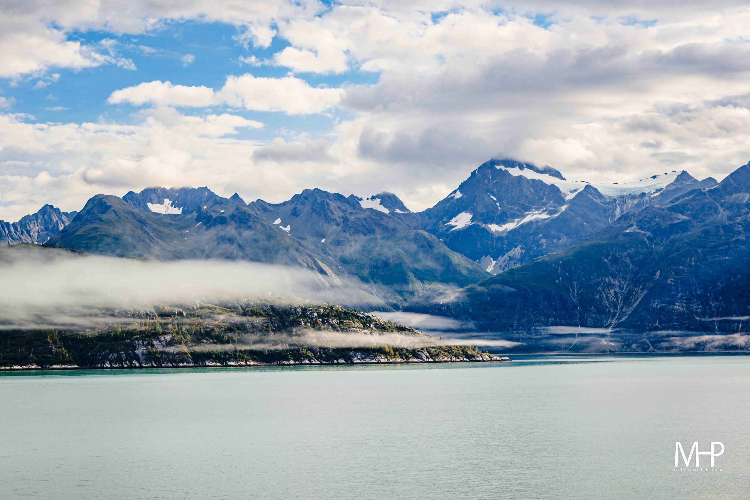 A mountain range with snow-capped peaks under a partly cloudy sky, located near a body of water with calm, greenish surface.