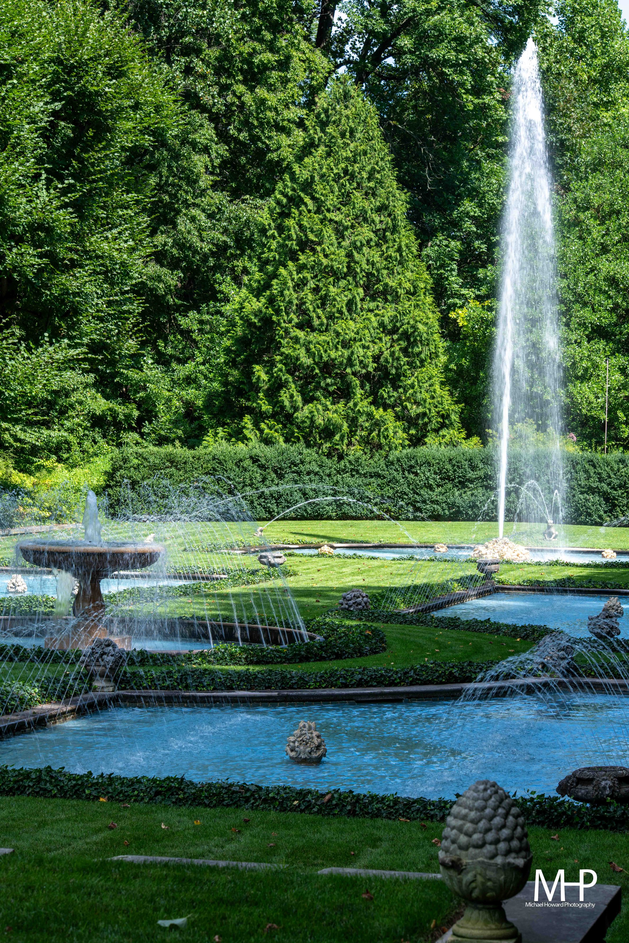 A lush, green park with a large fountain featuring multiple water jets and sculptures, surrounded by neatly trimmed grass and trees.