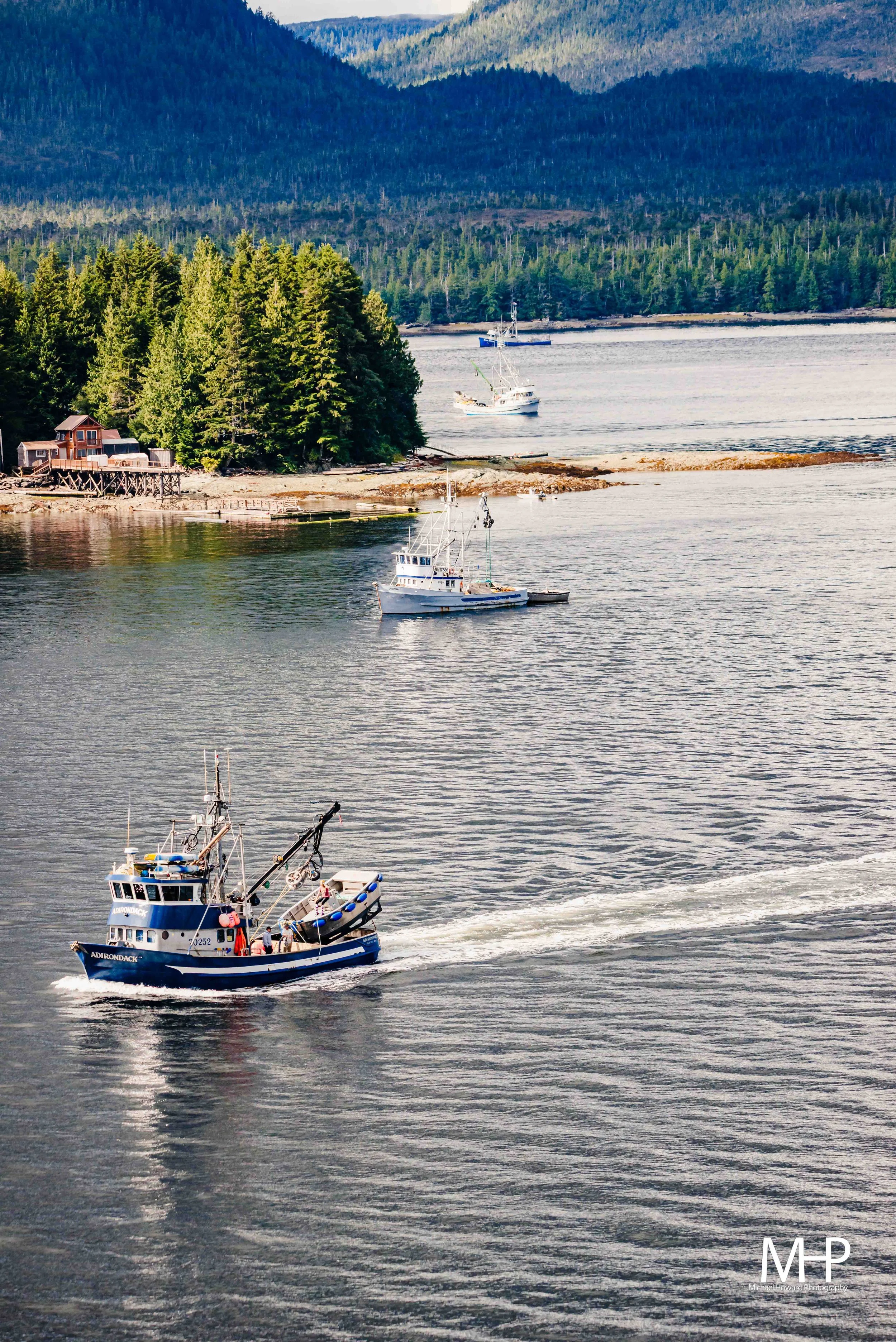 A black and blue boat in water with a smaller black boat attached, while three other boats are visible in the background near a wooded shoreline and distant mountains.