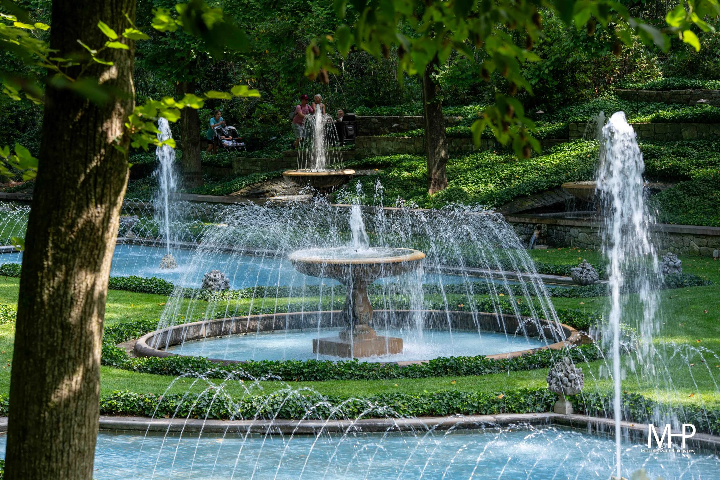 A park scene with multiple fountains in a pond, surrounded by greenery, with a group of people walking nearby.