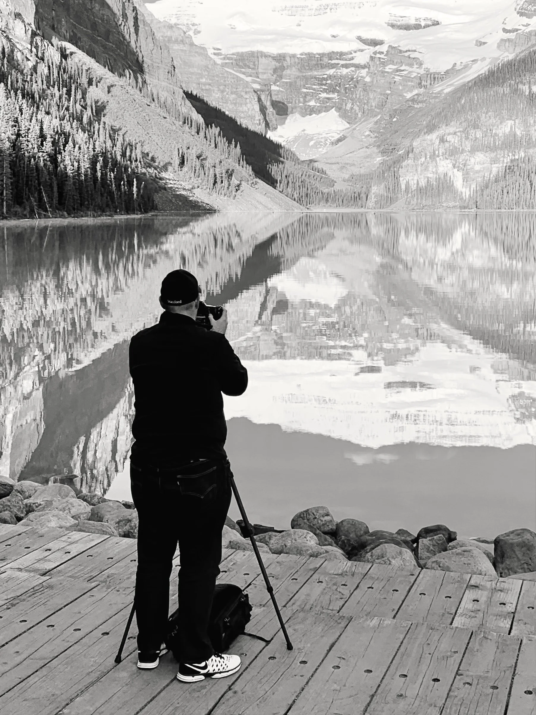 A person standing on a wooden dock, taking a photograph of a mountainous landscape with a large lake, snow-capped peaks, and trees in the background.