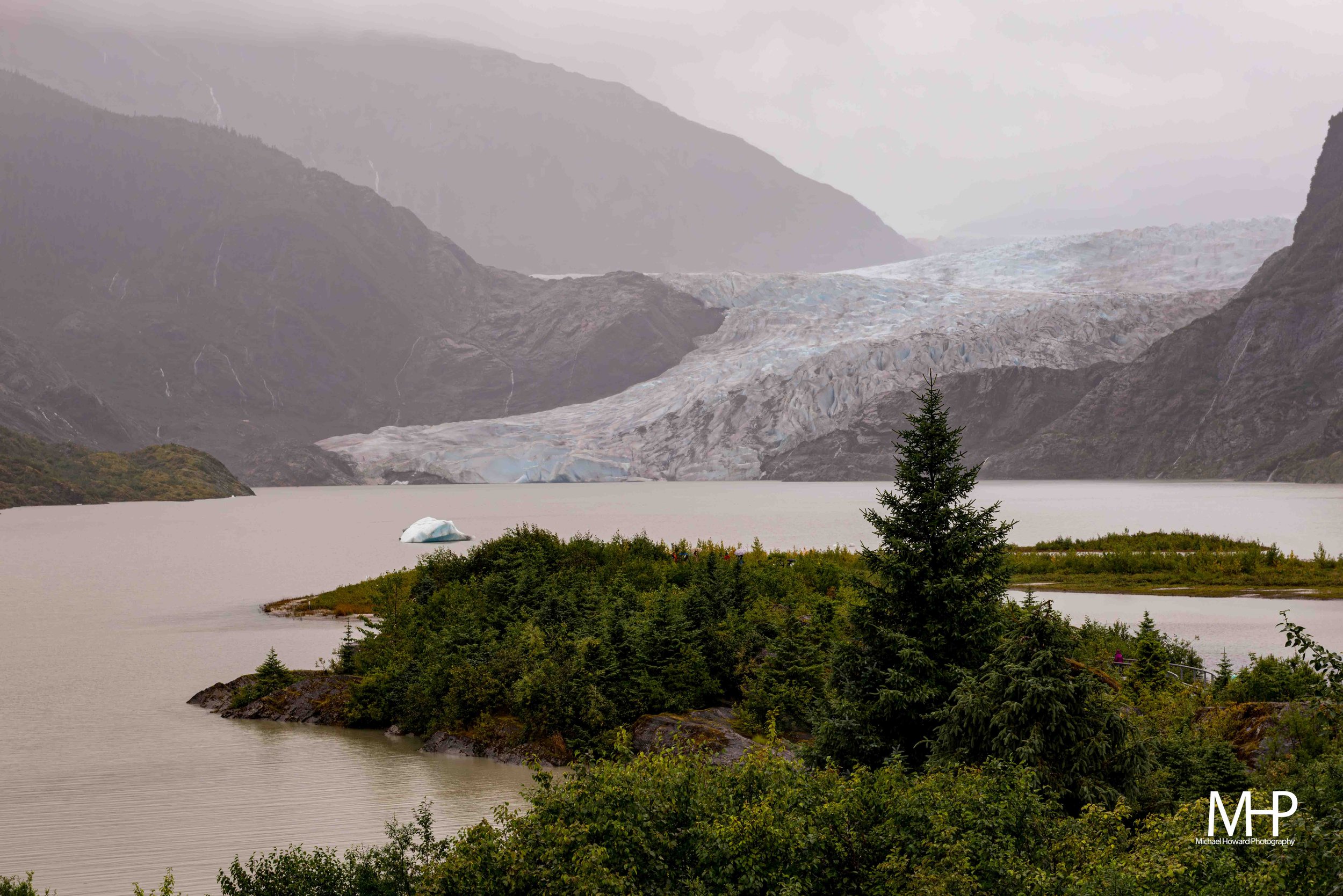 A scenic landscape featuring a large glacier flowing into a lake, surrounded by mountains and lush green trees in the foreground.