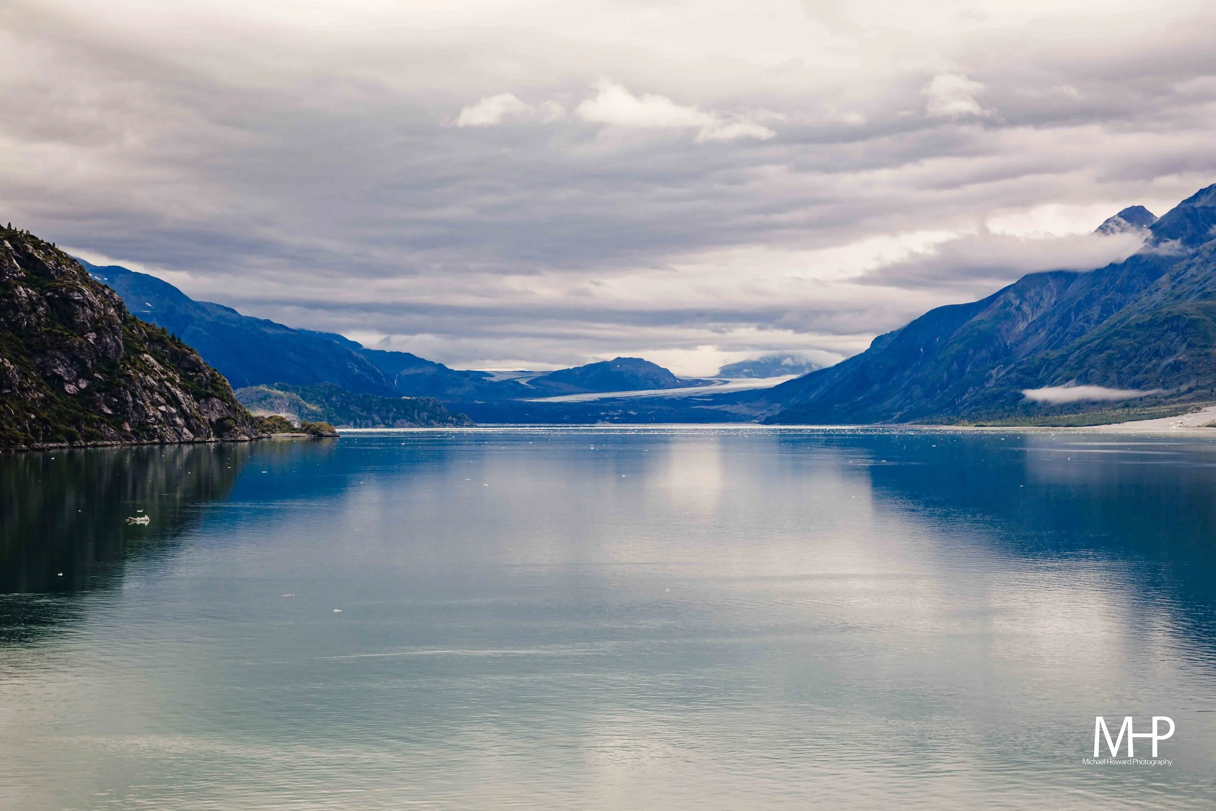 A wide river flowing through a mountainous landscape under cloudy sky, with green and rocky hills on both sides and mountains in the distance.