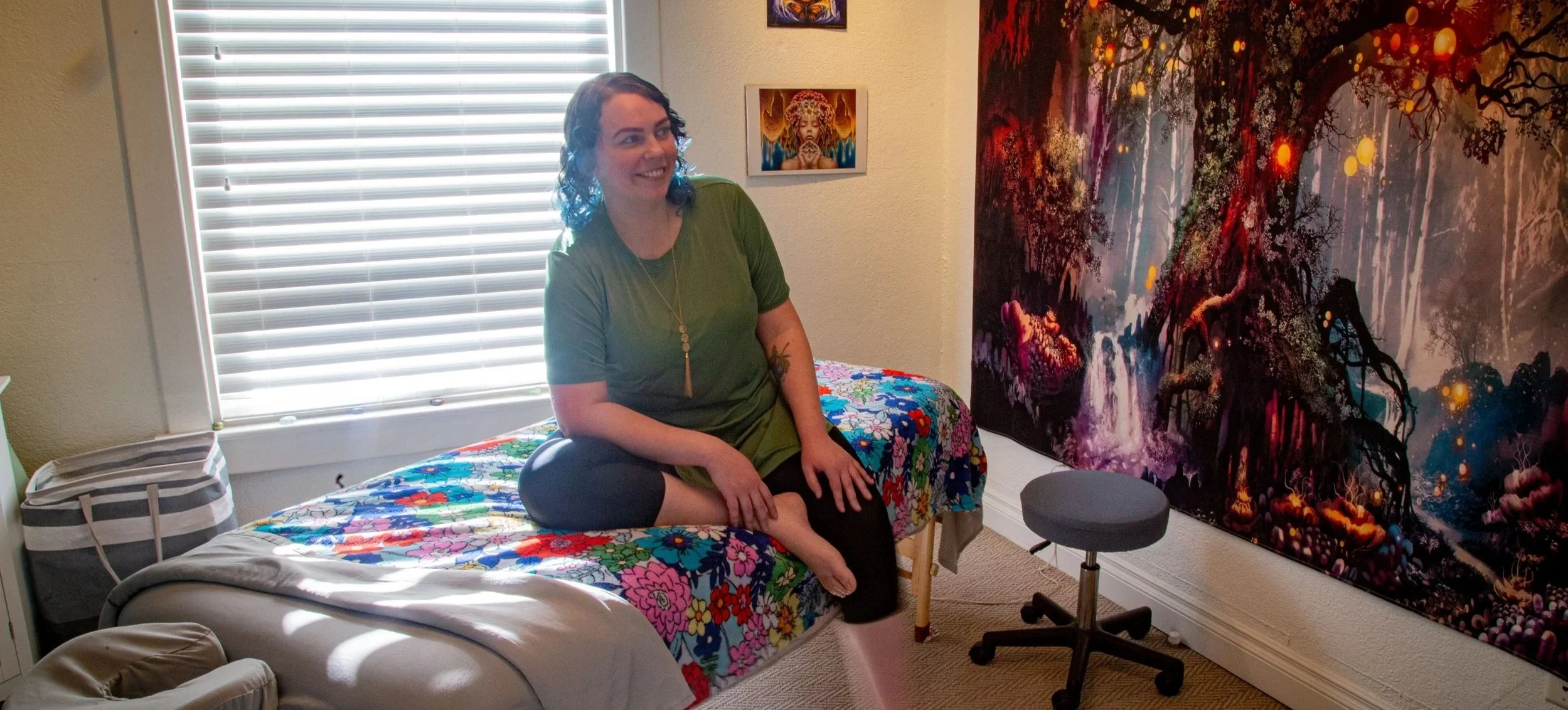 A woman with curly dark hair and a smile, sitting on a bed covered with a vibrant, multicolored floral blanket. She is dressed in a green top and black leggings, barefoot, and has a necklace with a pendant. The room has a window with closed blinds, colorful artwork on the wall, including a large fantasy-themed tapestry featuring a forest scene, a window, and small glowing orbs. There is also a gray stool and a striped bag on the bed.