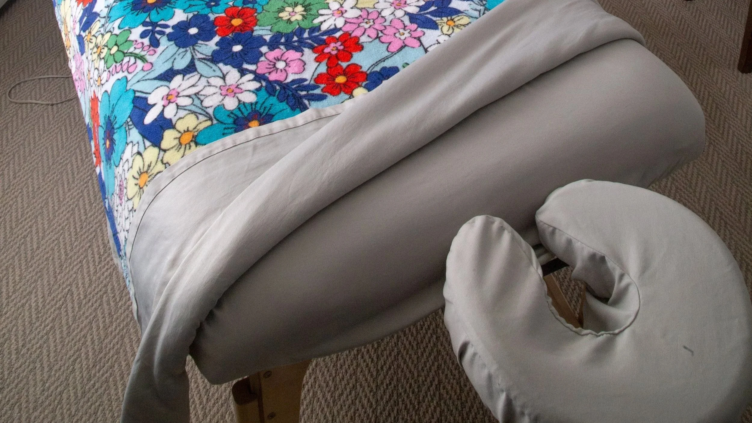 A massage table with a colorful floral comforter on brown patterned carpet in the clean and inviting treatment space of this south denver massage studio