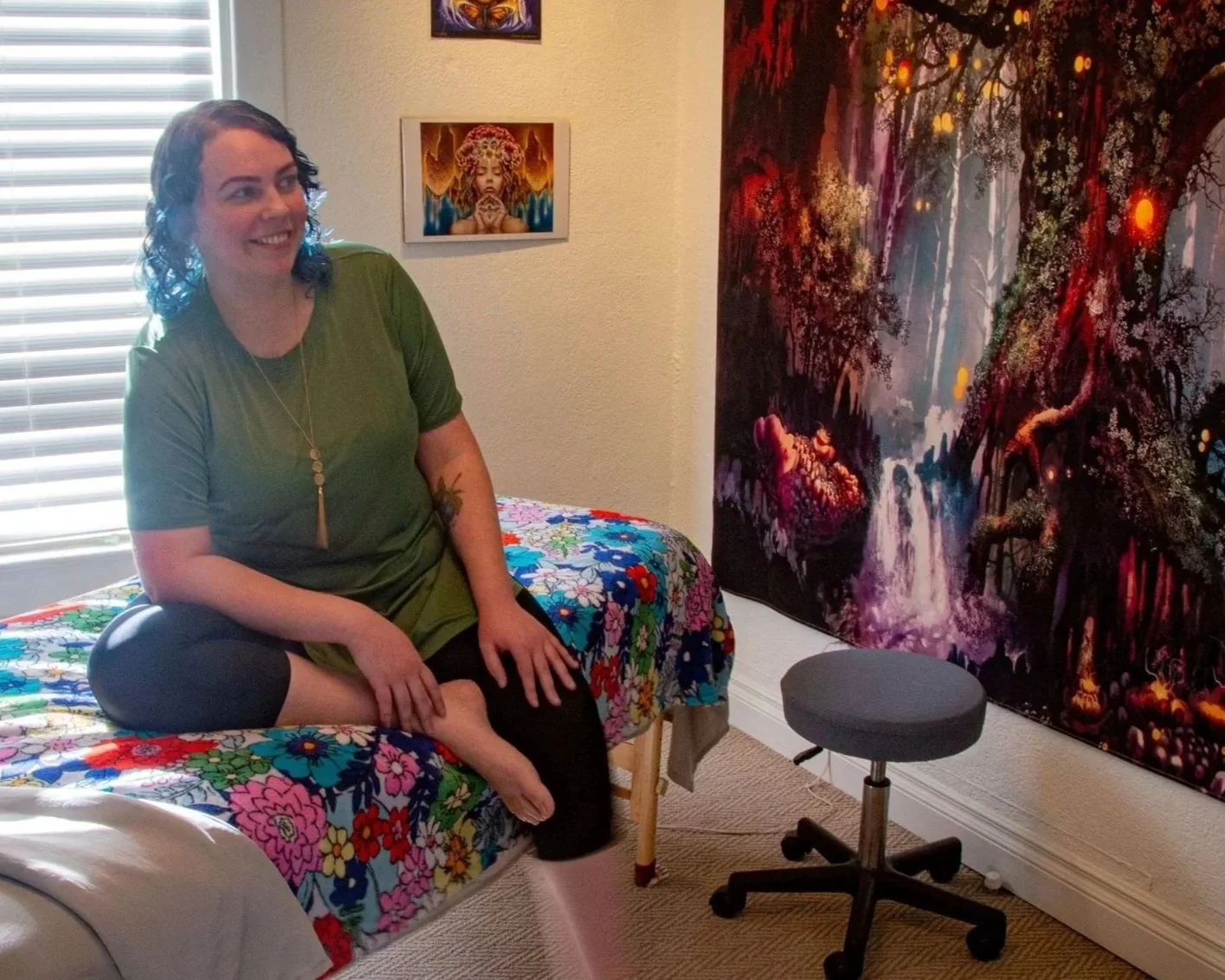 A woman with curly dark hair, wearing a green t-shirt and black leggings, sitting on a bed with a colorful floral bedspread. She is smiling and looking to her left. Behind her is a window with closed blinds, and on the wall are two pieces of artwork: one of an angelic figure with a crown of flowers and a serene face, and another smaller painting. To the right of the woman, there is a large, vibrant, fantasy-style painting of a forest with waterfalls and glowing lights, and a gray rolling stool on wheels.