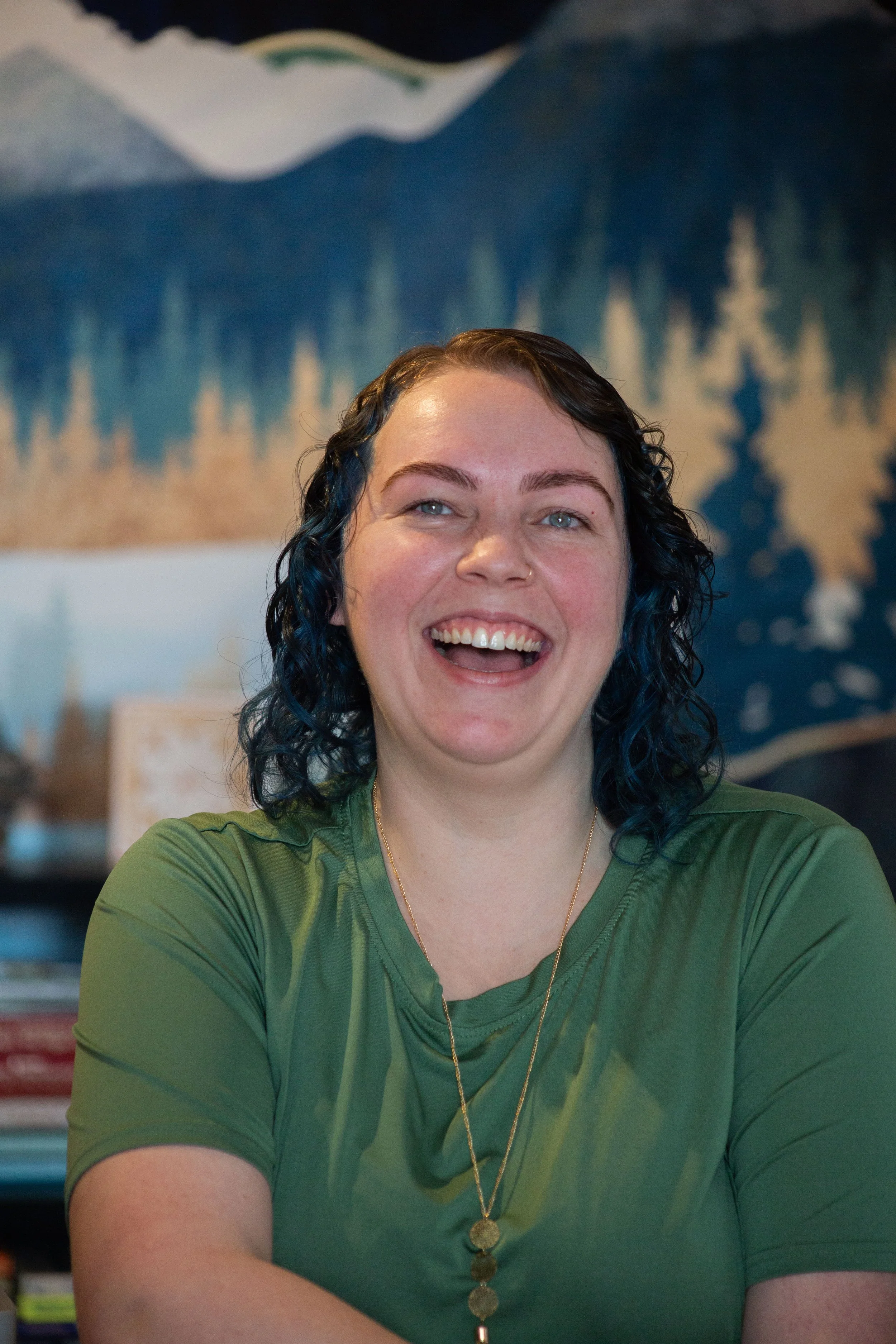 Aspen, licensed massage therapist and owner of Odyssey,  woman with curly dark hair and light skin, smiling and wearing a green shirt and a gold necklace with coin pendants. The background shows an abstract mural or painting of a mountain landscape.