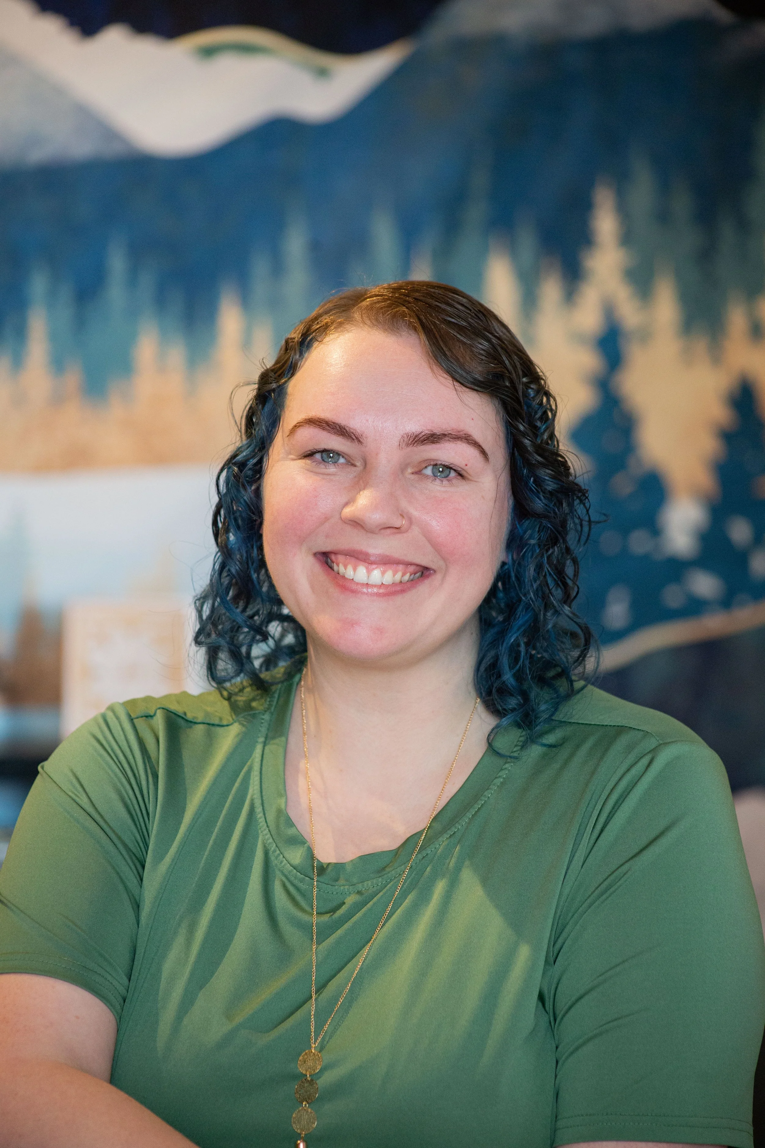 Aspen, licensed massage therapist of Odyssey holistics, is a smiling woman with blue wavy hair, wearing a green shirt and a long gold necklace, sitting in front of a mountain landscape painting.