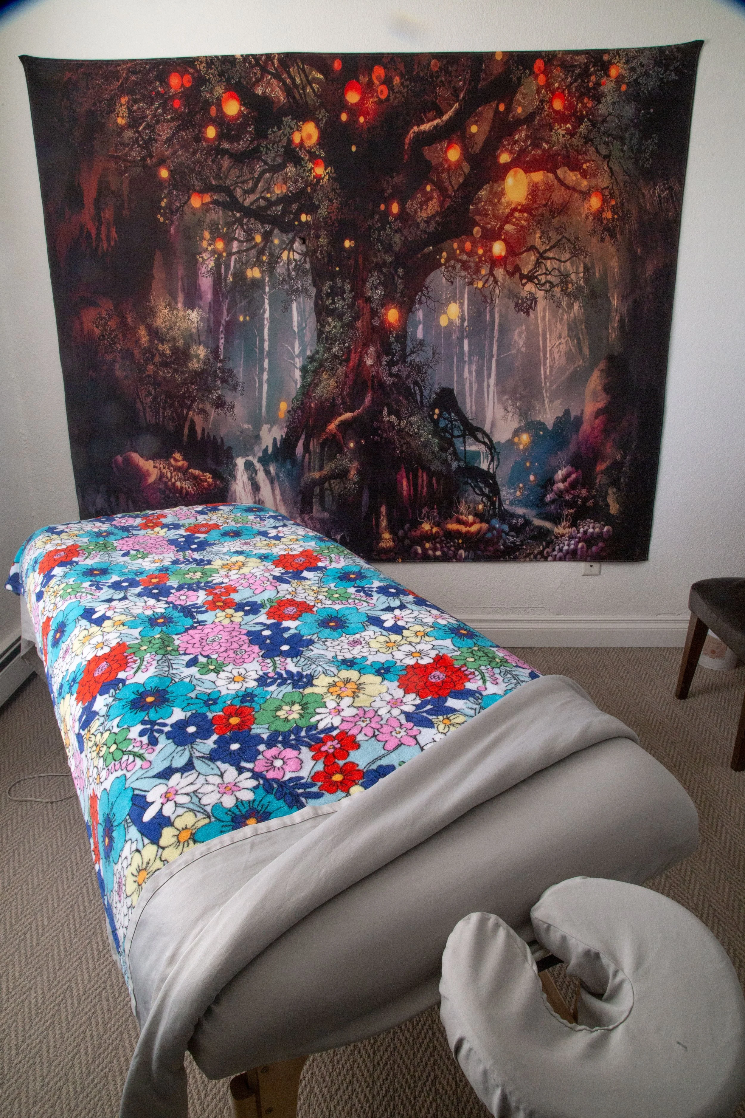 A massage table covered with a colorful floral blanket in a clean and inviting massage therapy room, with a large tapestry of a dark enchanted forest scene hanging on the wall behind it.