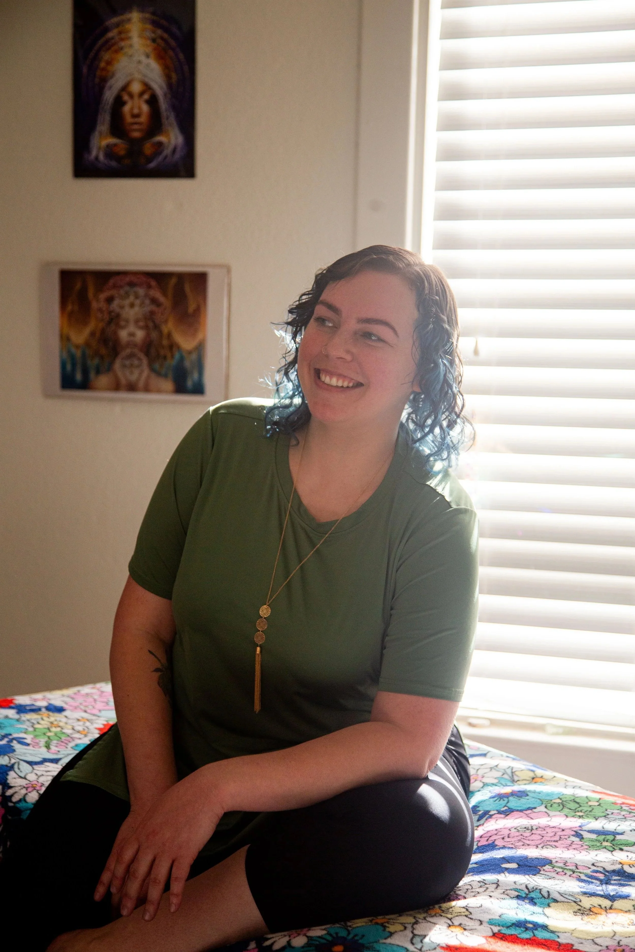 candid photo of Aspen, therapist at Odyssey massage studio, sitting on her table, smiling serenely into the distance. She is wearing a green cotton tee, and has loosely curled brown hair