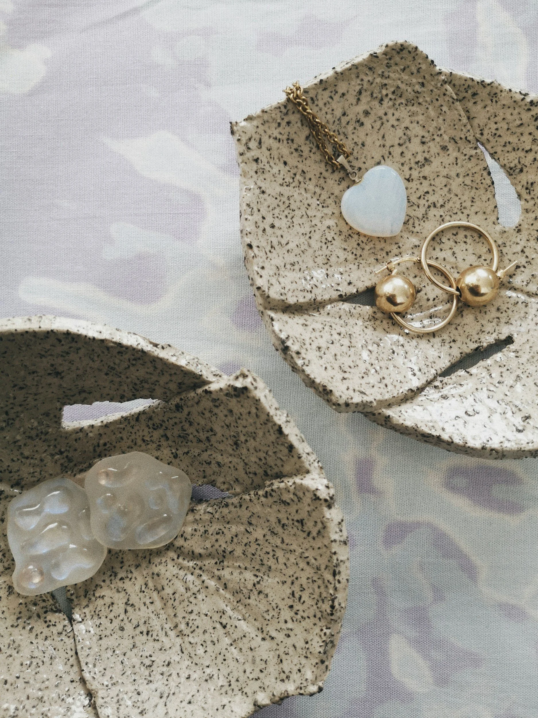 Two speckled, textured ceramic dishes, each holding jewelry: one with a white heart-shaped pendant on a gold chain, and the other with gold hoop earrings featuring large pearl accents, on a light-colored fabric surface.