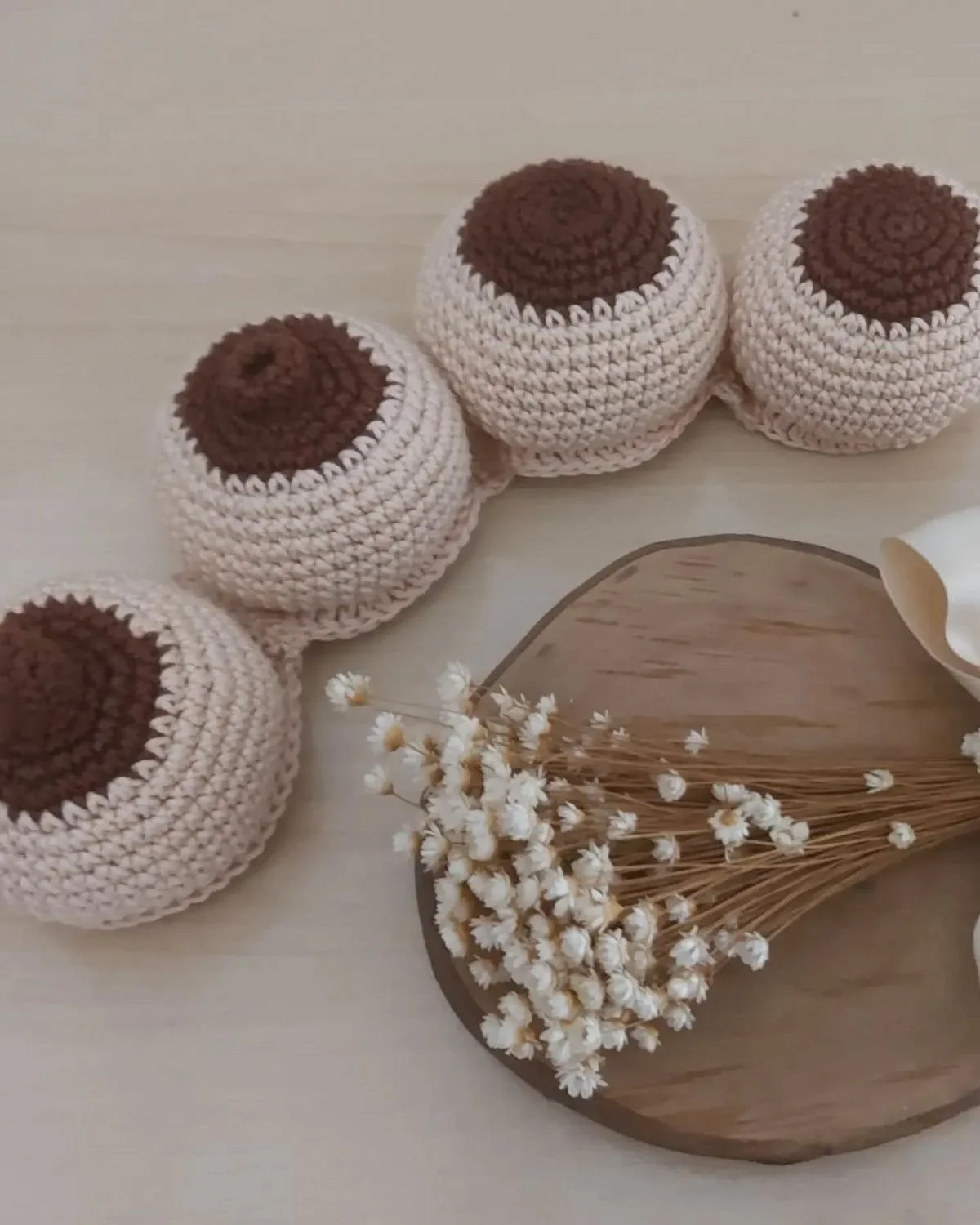 Five crocheted round baskets with brown tops are arranged in a line on a light-colored surface. To the right, a wooden tray holds a bunch of small white flowers with brown stems, next to a white rolled paper or napkin.