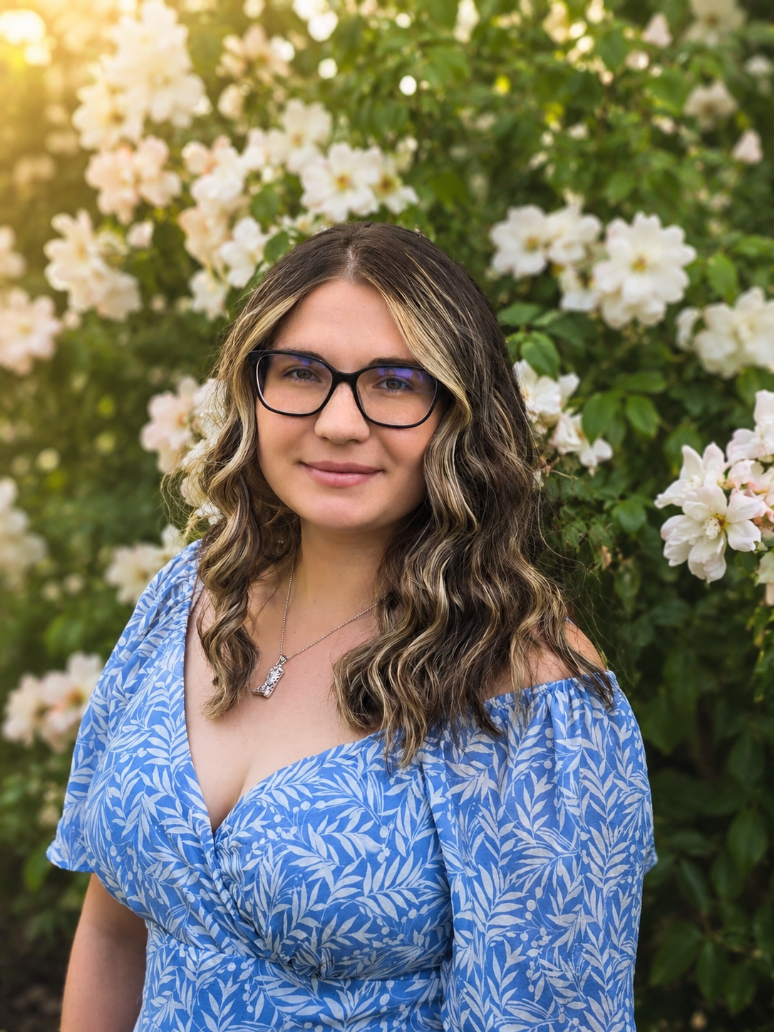A woman with wavy brown hair wearing glasses, a blue leaf-patterned dress, and a necklace with a small pendant, standing outdoors in front of blooming white flowers.