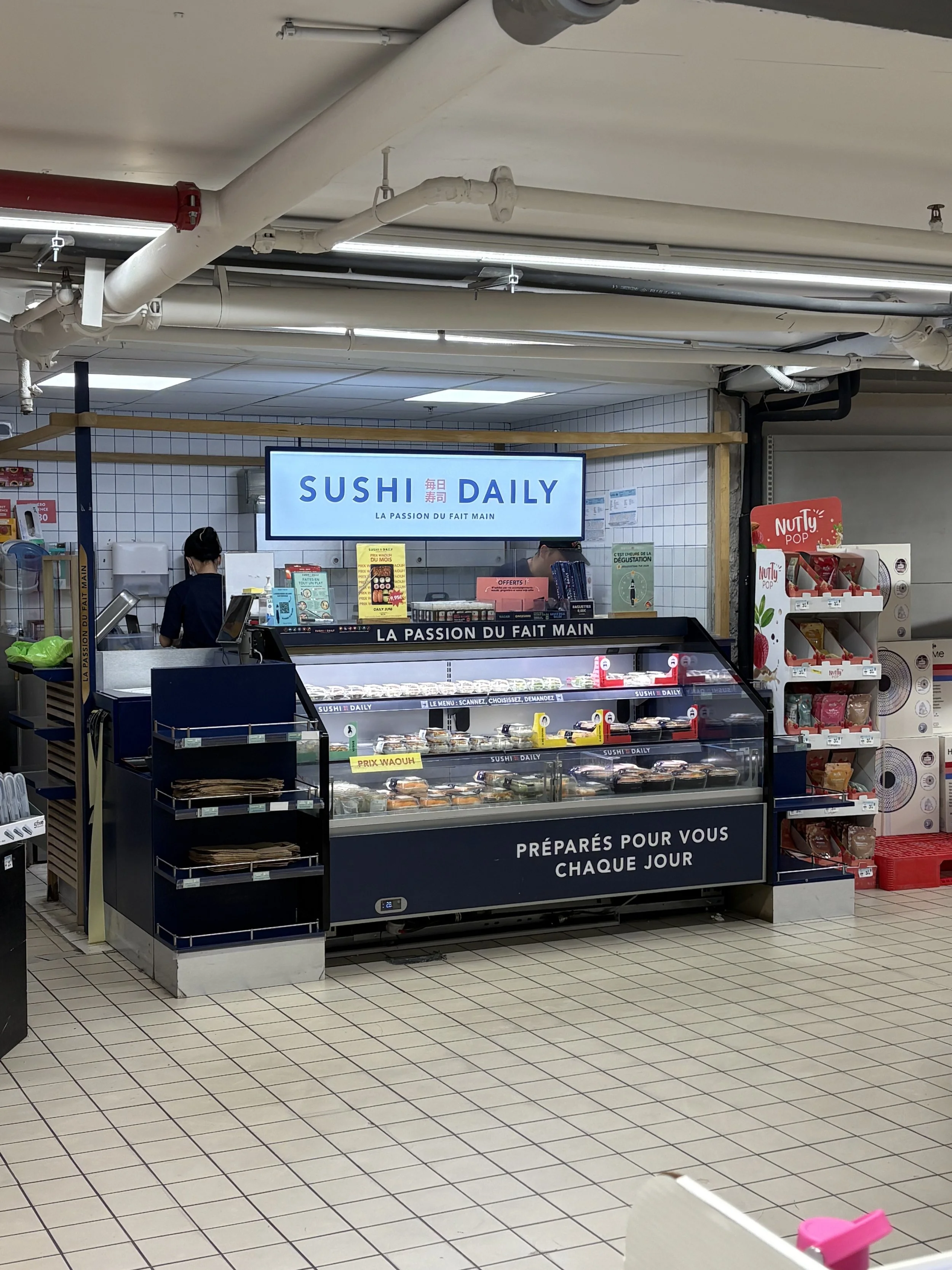 Un comptoir de vente en supermarché avec des plats préparés pour sushi, une enseigne lumineuse blanche indiquant 'SUSHI DAILY' et la phrase 'LA PASSION DU FAIT MAIN'. Sur le côté, une étagère avec des snacks et des produits. Le sol est en carrelage blanc avec des lignes noires.