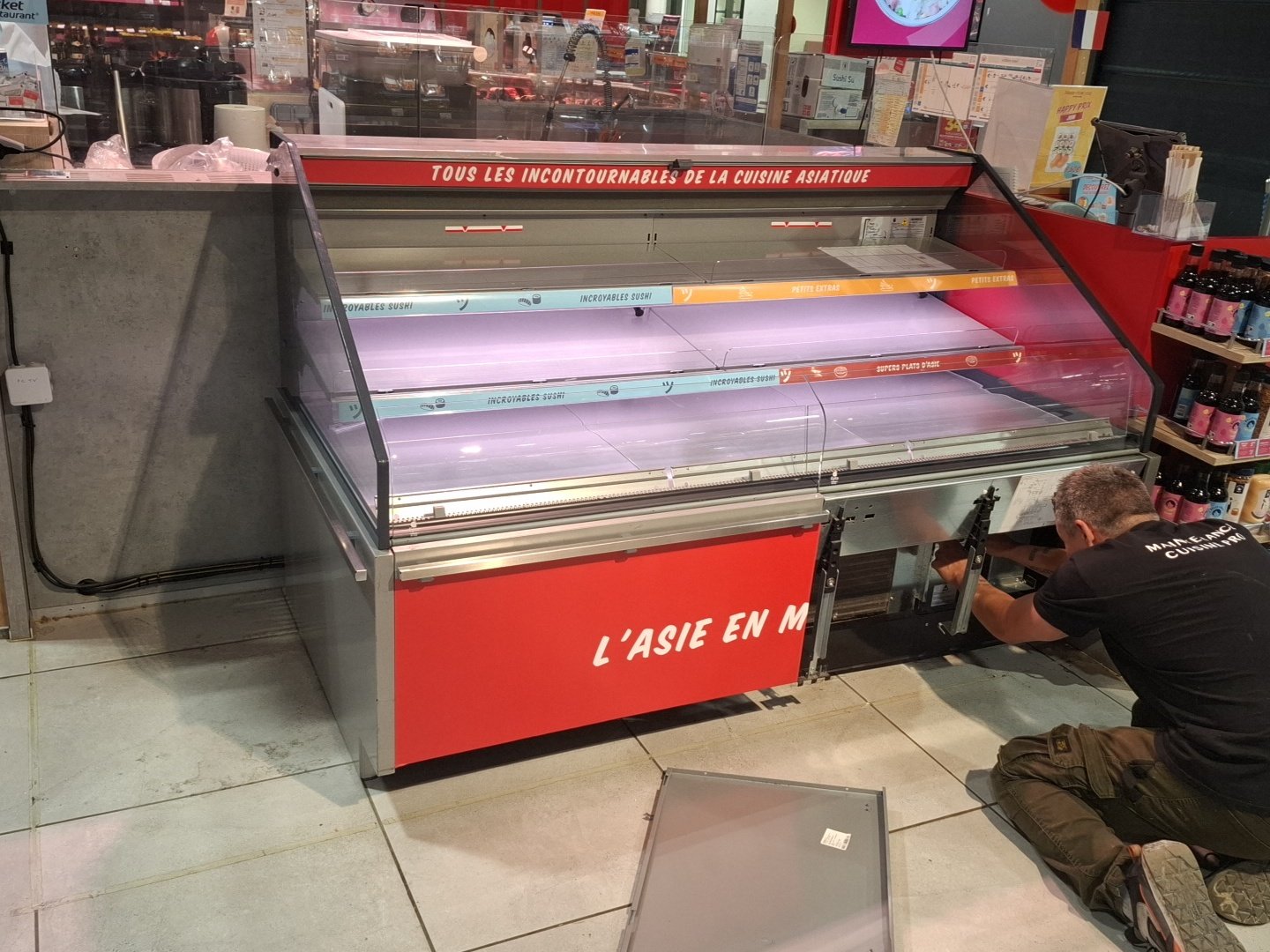Un homme travaille à la réparation d'un grill de vitrine de sushi vide dans un supermarché, situé dans une zone d'affichage de produits, avec des étagères de bouteilles à proximité.