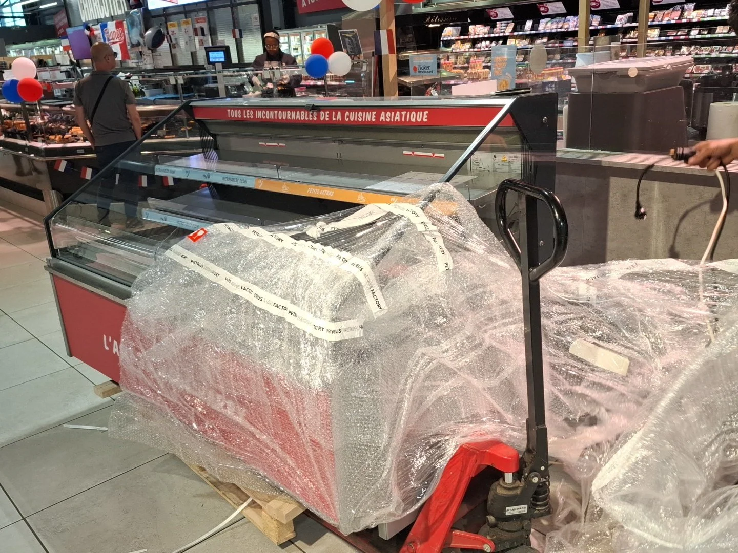 Vitrine réfrigérée en déballage, entourée de plastique à bulles, dans une supérette. Deux personnes dans le fond, une caisse enregistreuse, décorations de ballons, offre mentionnant la cuisine asiatique.