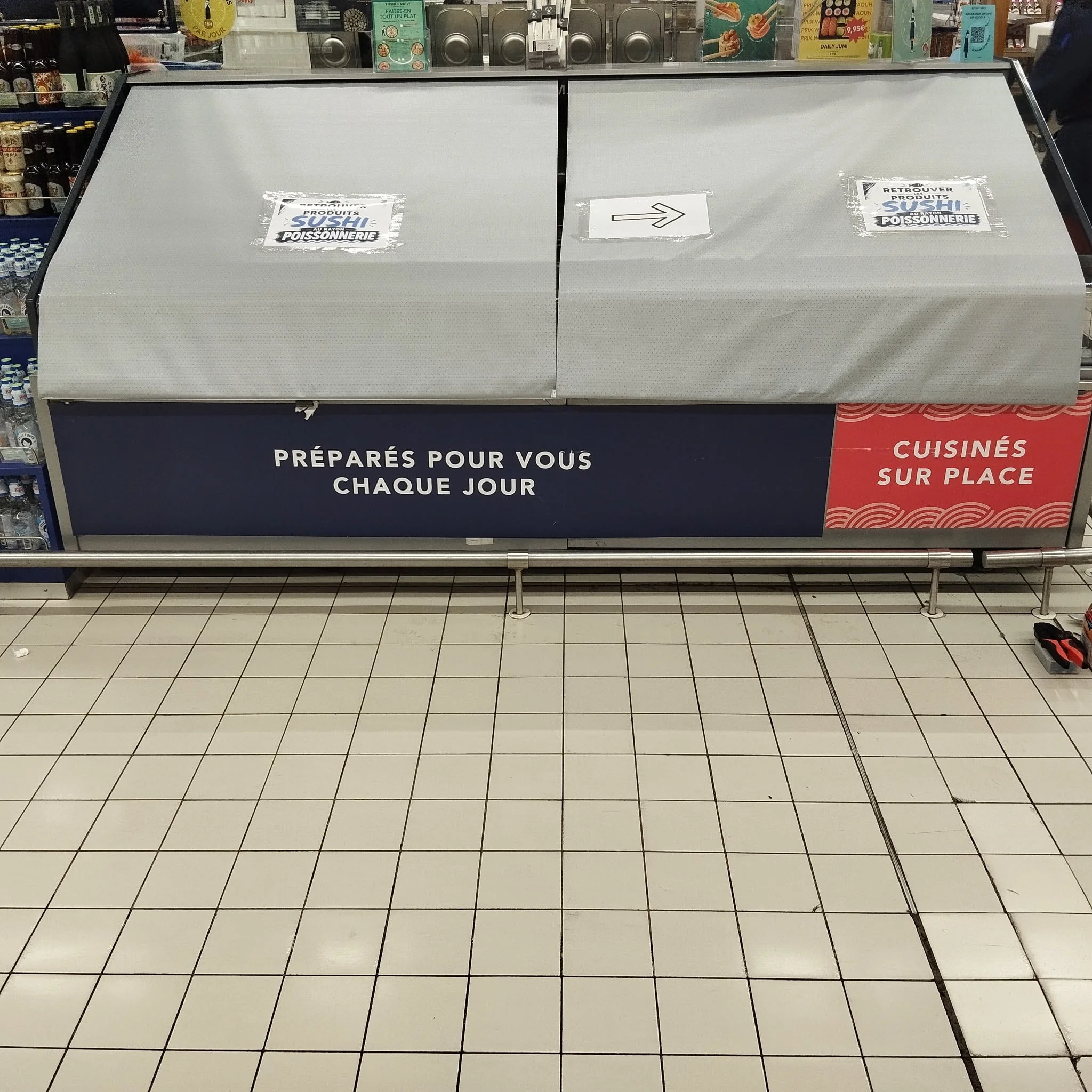 Une affiche sur un présentoir de supermarché avec un grand panneau blanc, deux étiquettes en papier avec une flèche et des textes en français, mentionnant VENTE EN SOUS-POISSONNIER et RÉTROUVÉ PRODUITS SOUS-POISSON. La partie inférieure du panneau a une partie rouge avec le mot CUISINÉ SUR PLACE.