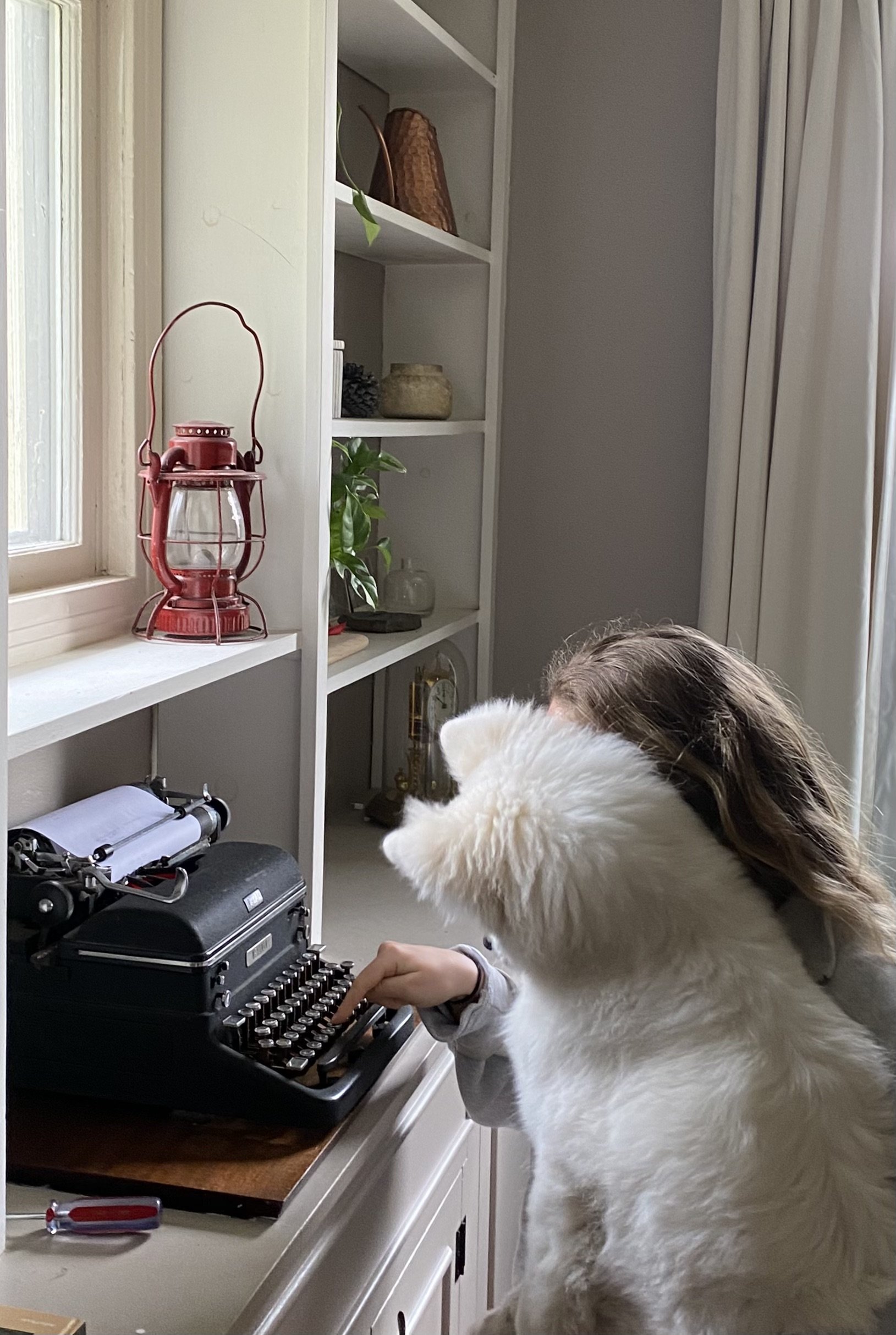 A child with light brown hair is sitting at a table using a vintage black typewriter. A large white fluffy dog, possibly a Samoyed, stands close beside the child, observing the typewriter. The setting appears to be a cozy room with built-in shelves displaying various decorative items like a red lantern, a planter with a green plant, and other trinkets. A window with white trim is on the left side of the image, allowing natural light into the room.