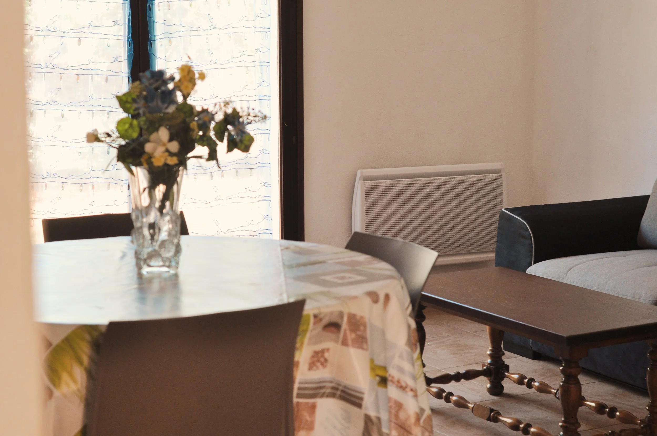 Une table recouverte d'une nappe à motifs, avec un vase contenant des fleurs floues au premier plan. En arrière-plan, une fenêtre avec des rideaux légers et un radiateur dans un salon ou une salle à manger.