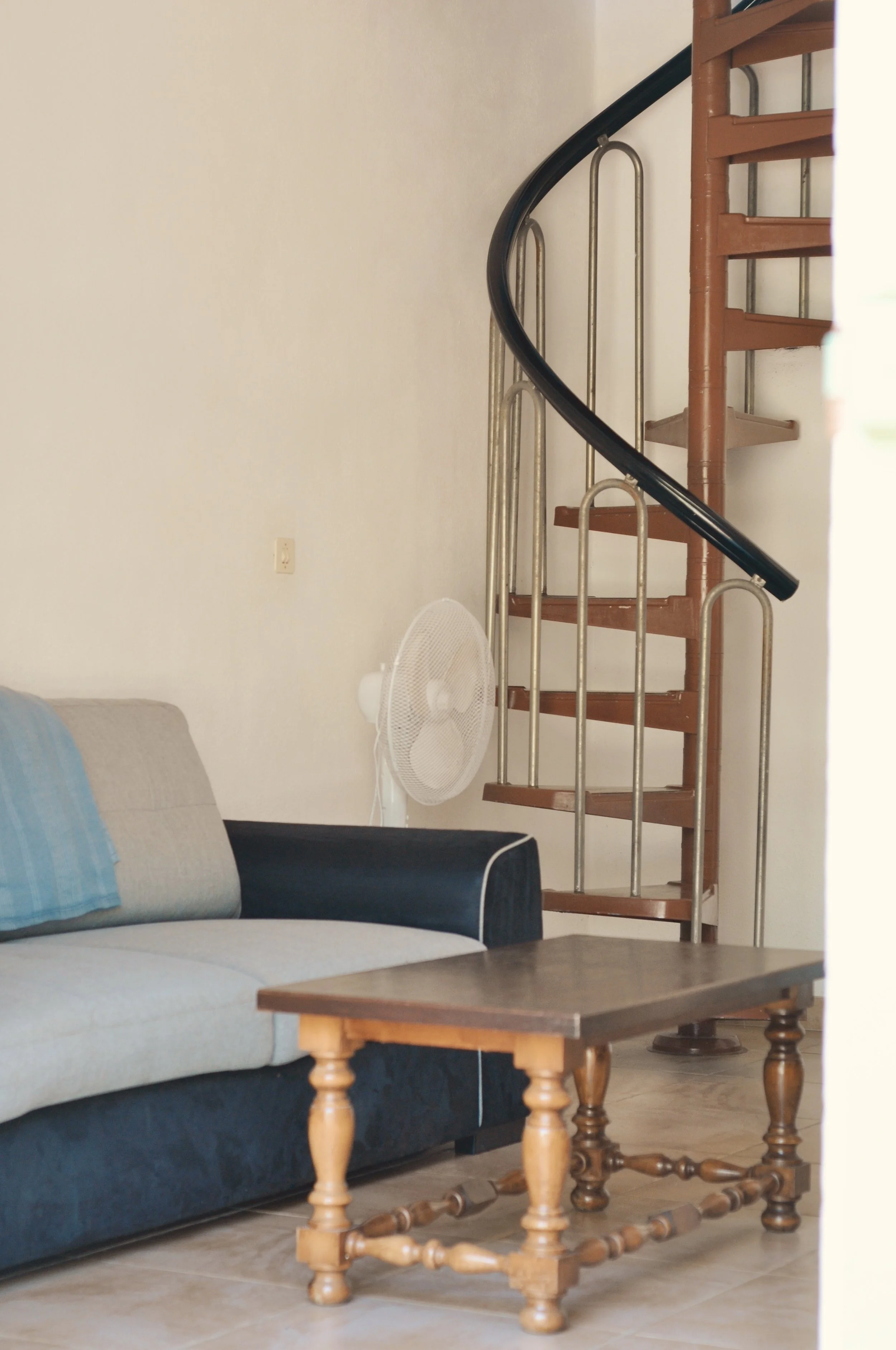 Salon avec un canapé bleu et une table en bois, un ventilateur blanc, un escalier en bois à spirale, et un mur beige avec une prise électrique.