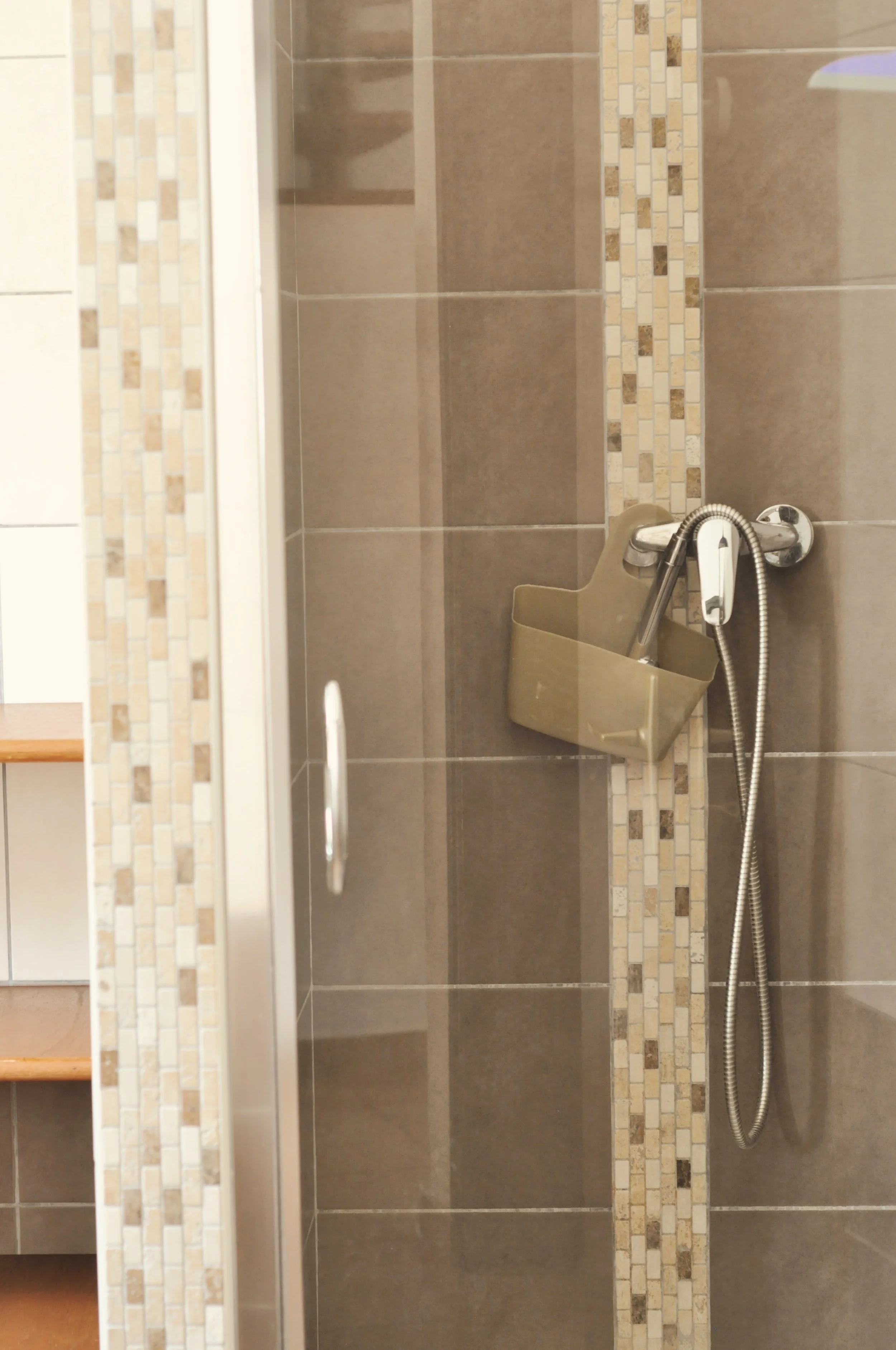 Une salle de douche avec des carreaux beige et une bande de mosaïque. Il y a une douchette avec un tuyau flexible et un porte-rasoir ou une petite étagère en plastique beige fixée au mur.