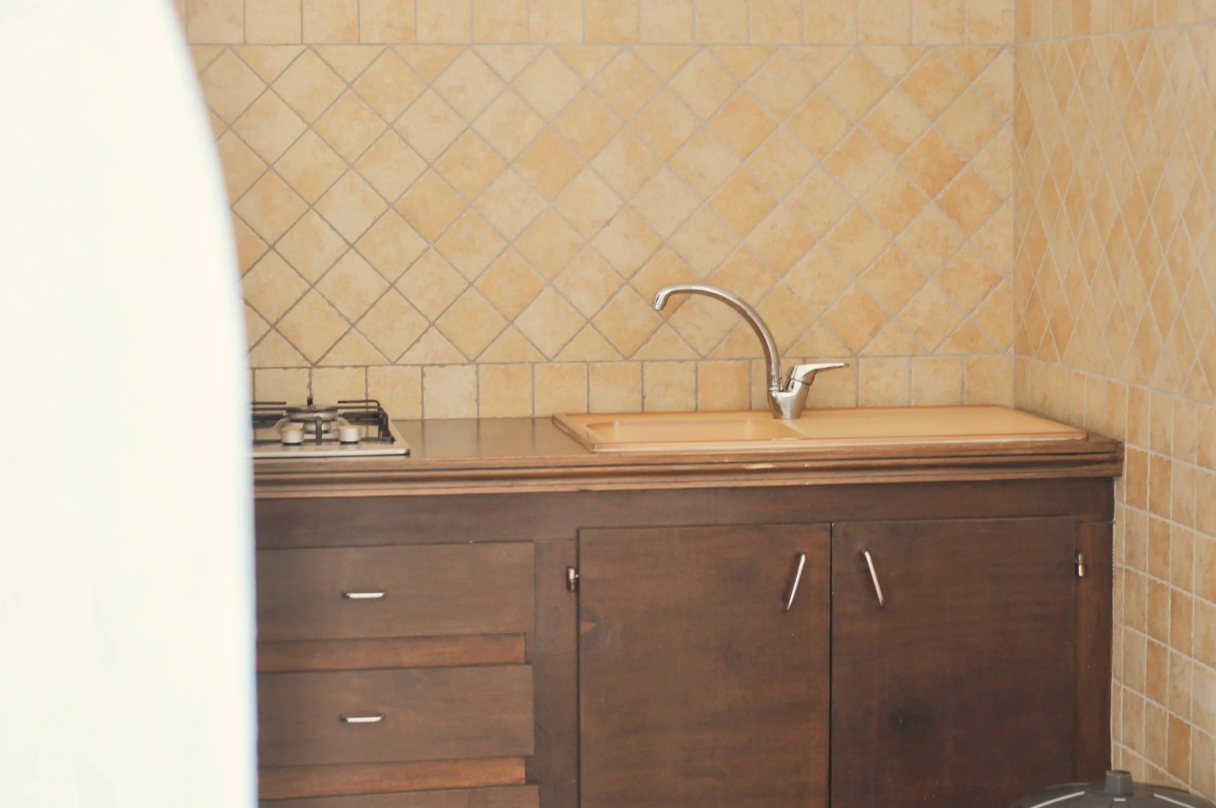 Une cuisine avec un plan de travail en bois, un évier en dessous d'un robinet en métal, une plaque de cuisson à gaz à gauche, des armoires en bois foncé en dessous, et des murs en carrelage beige avec motif en damier.