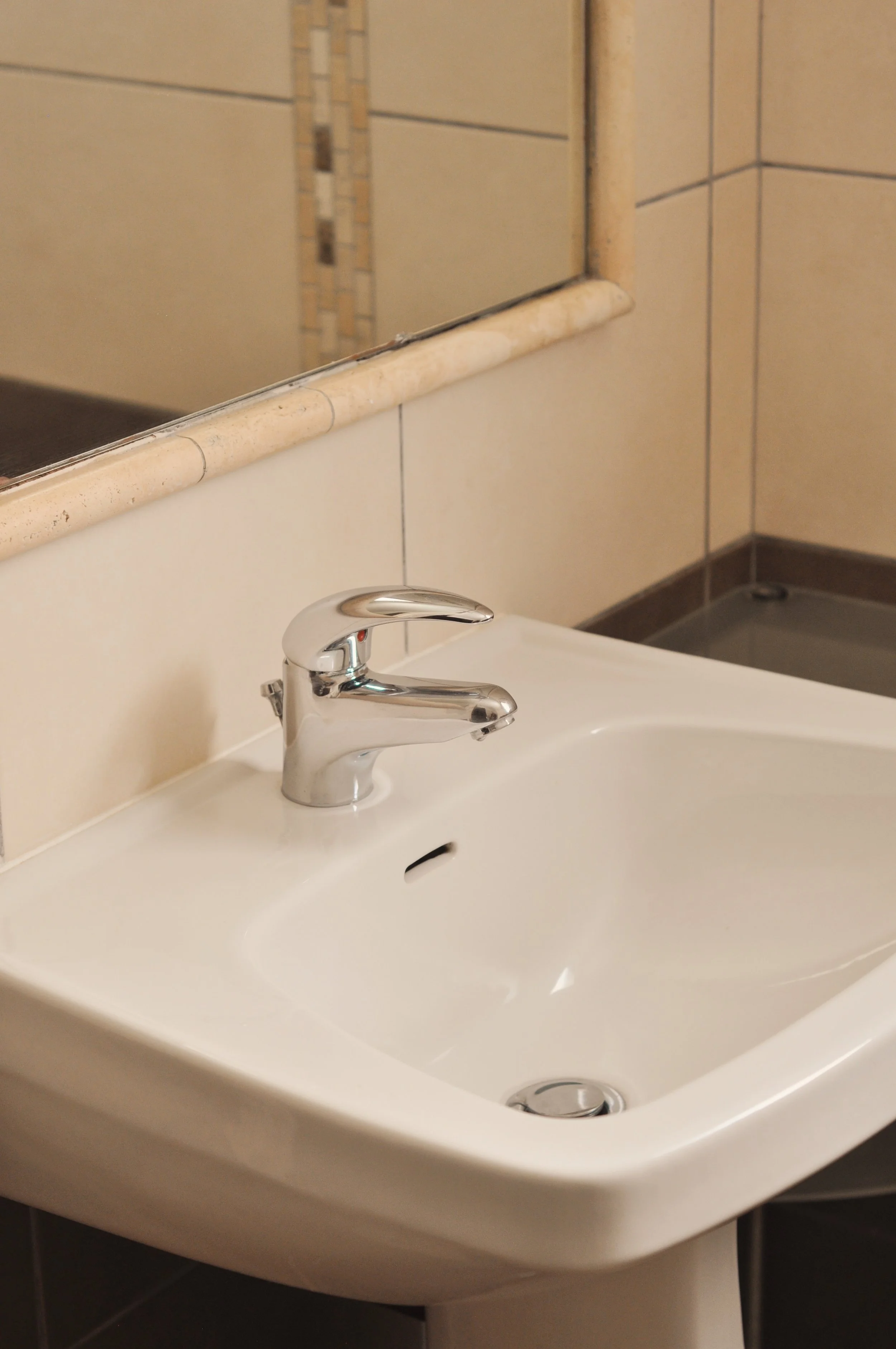 L'image montre un lavabo blanc avec un robinet en métal brillant dans une salle de bain, un grand miroir au-dessus, un mur carrelé beige avec une bande décorative, et un sol en carreaux sombres.