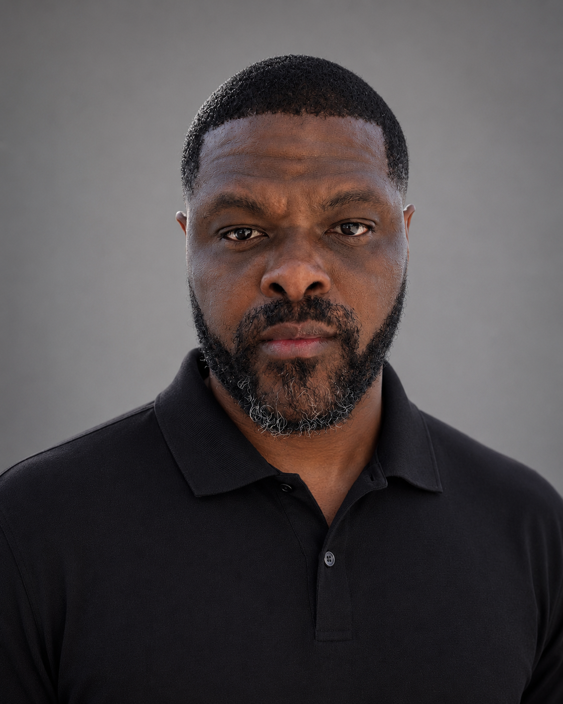 Close-up portrait of a serious Black man with a beard, wearing a black polo shirt, against a neutral gray background.