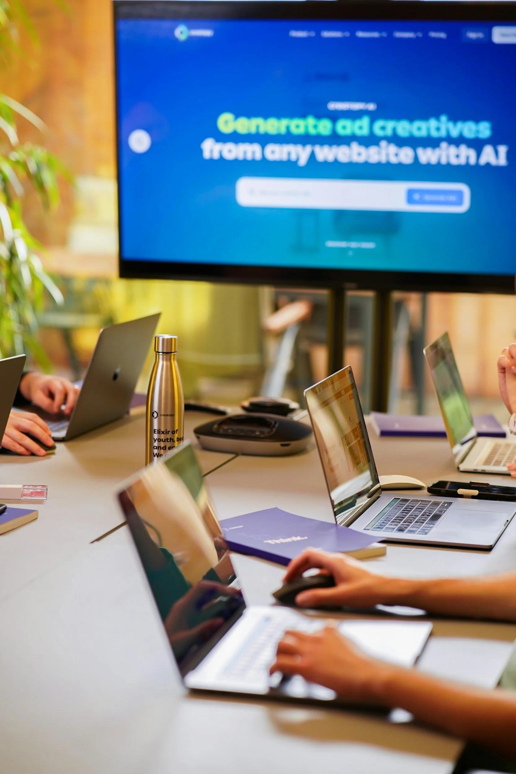 A conference room with people working on laptops around a table. A large screen displays a website with the text "Generate ad creatives from any website with AI."
