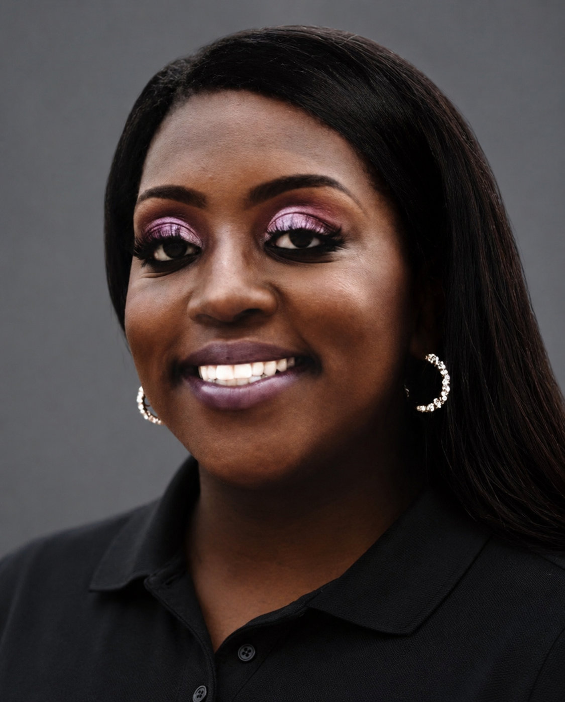 Close-up portrait of a woman with dark hair, wearing makeup with pink eyeshadow and lipstick, hoop earrings, and a black shirt with a collar, smiling against a gray background.