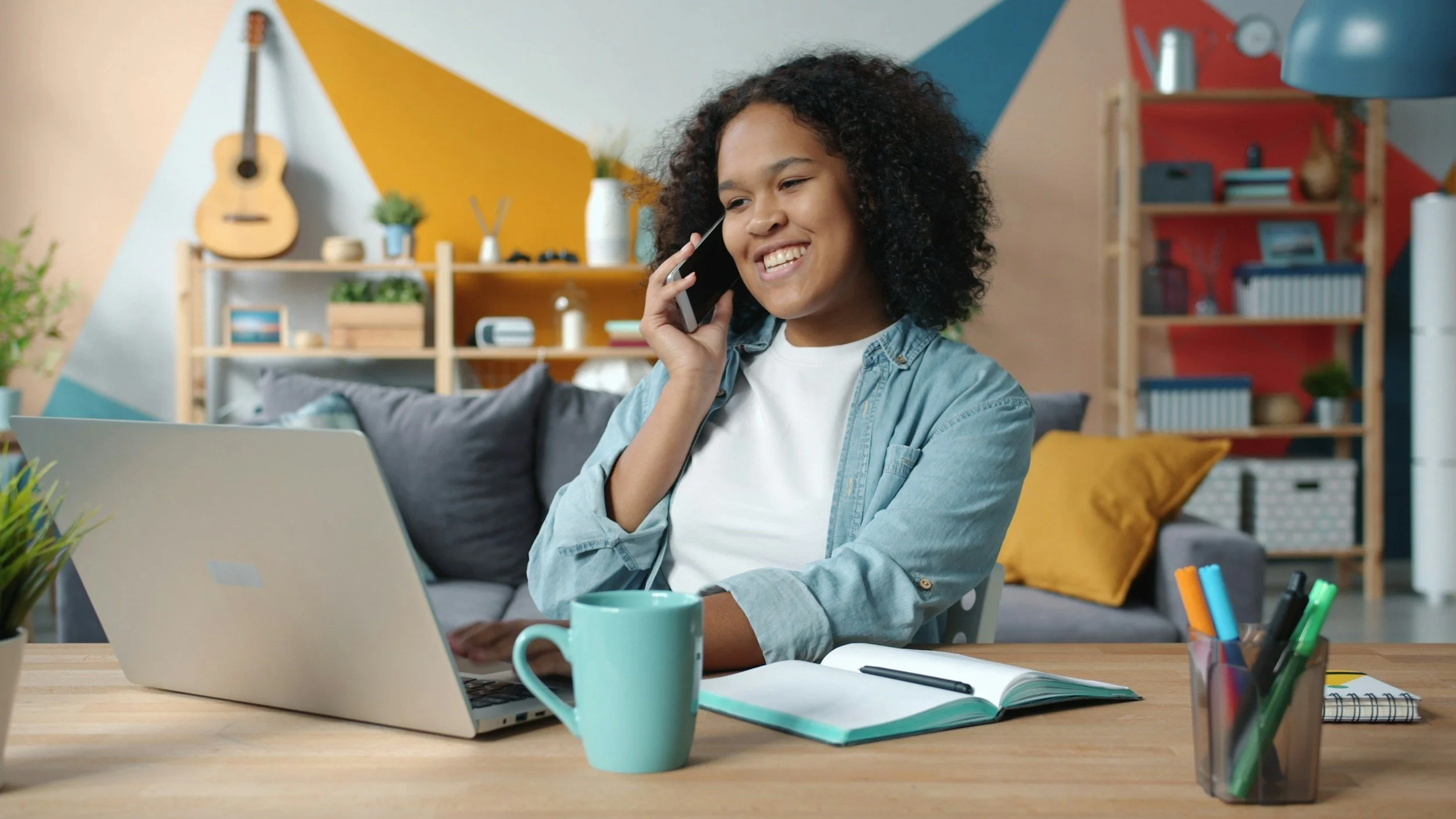 A young woman sitting at a desk with a laptop, talking on a cellphone, surrounded by notebooks, pens, and a mug, in a colorful and cozy living room.