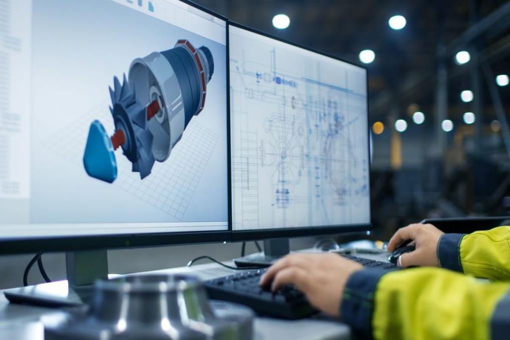 Person working on dual monitors displaying 3D CAD model and technical drawings in an industrial or engineering setting.