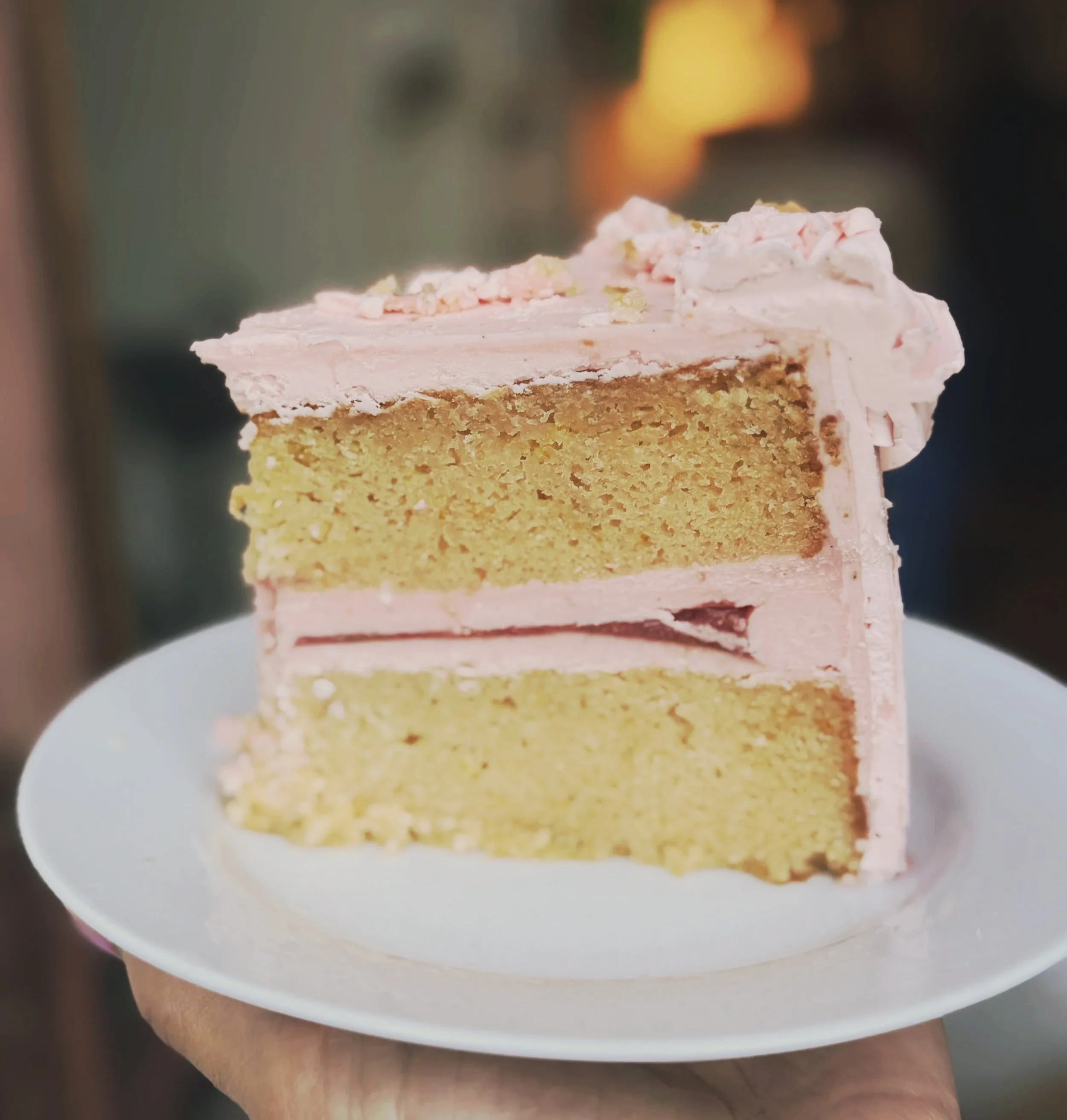 A slice of layered cake on a white plate, with pink and white frosting and small decorative topping, on a wooden surface.