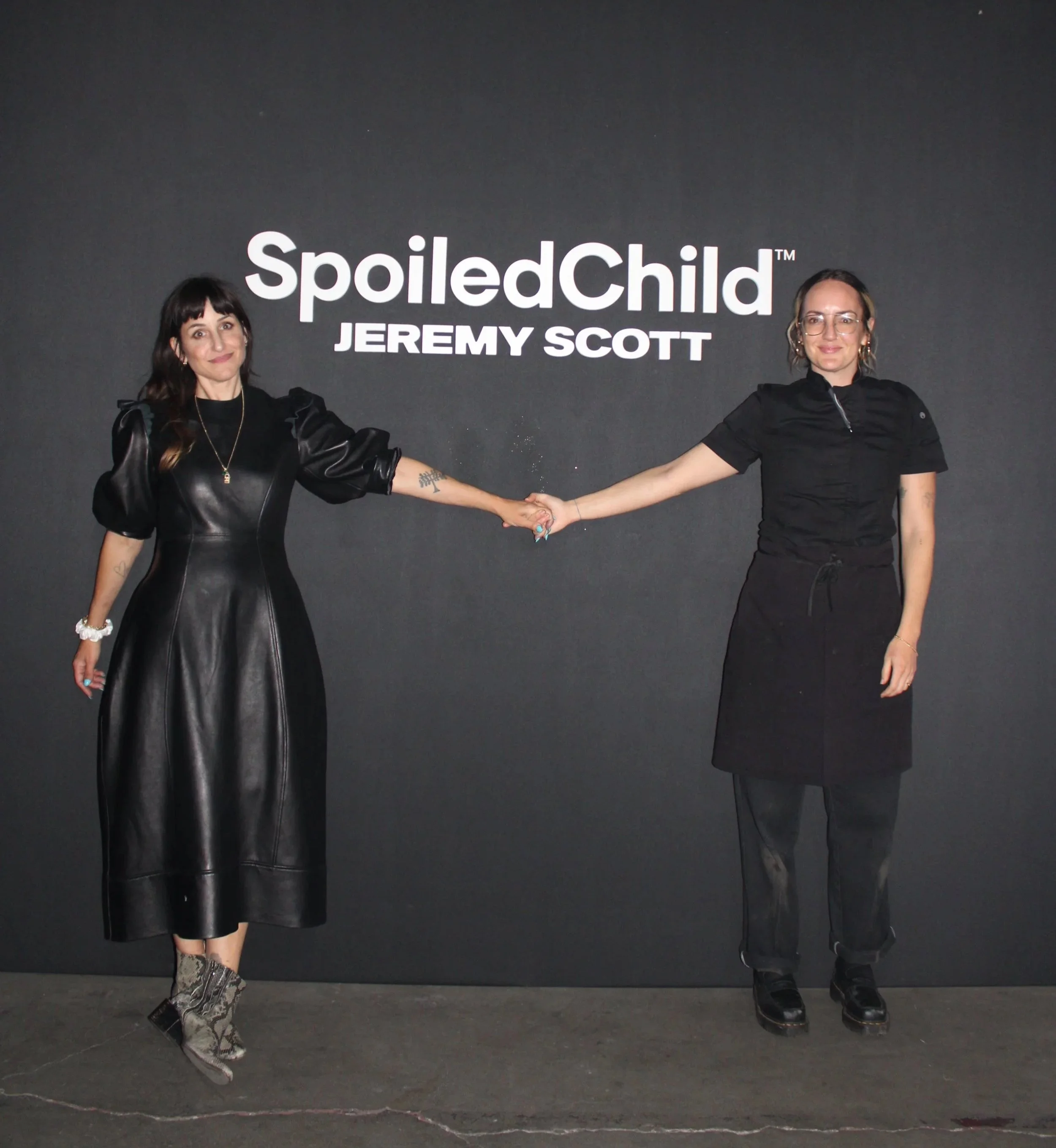 Two women standing in front of a dark background with the words 'SpoiledChild™ JEREMY SCOTT' displayed behind them. They are holding hands and smiling at the camera.