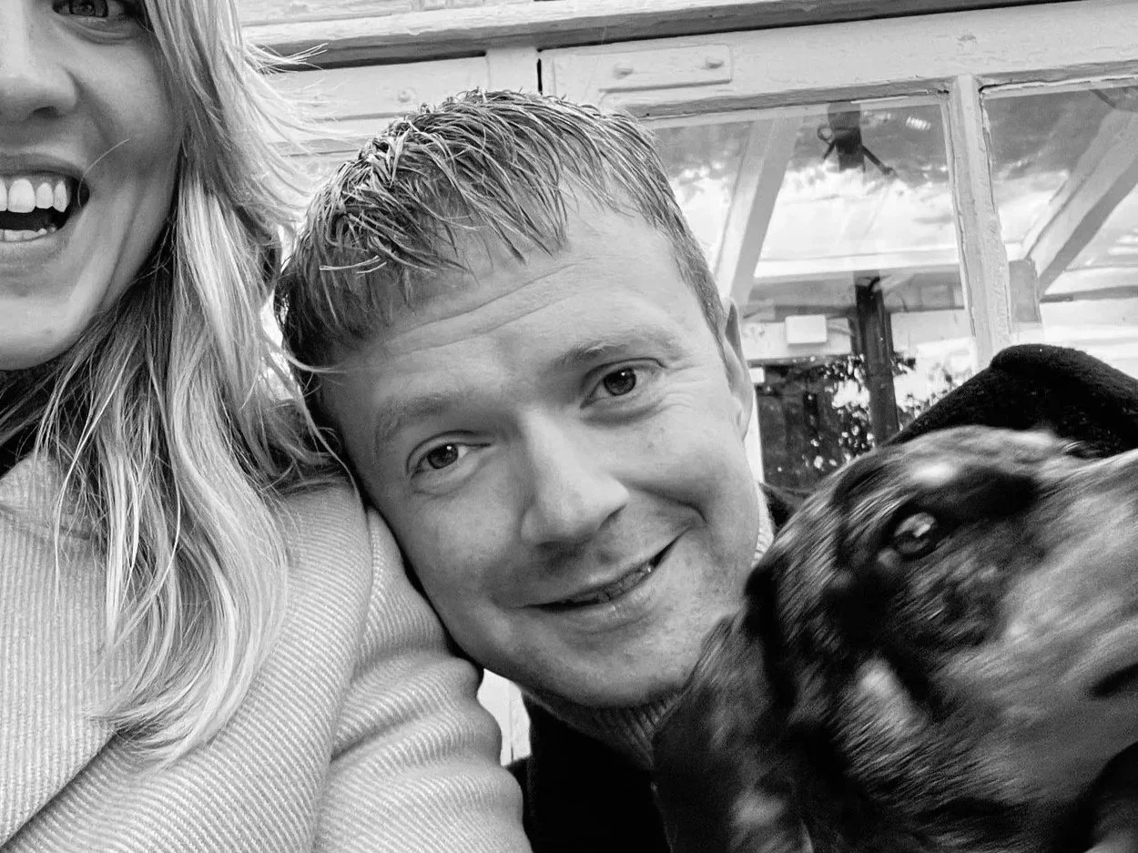 To personer, en mand og en kvinde, sammen med en hund, poserer for et foto i et hyggeligt miljø.