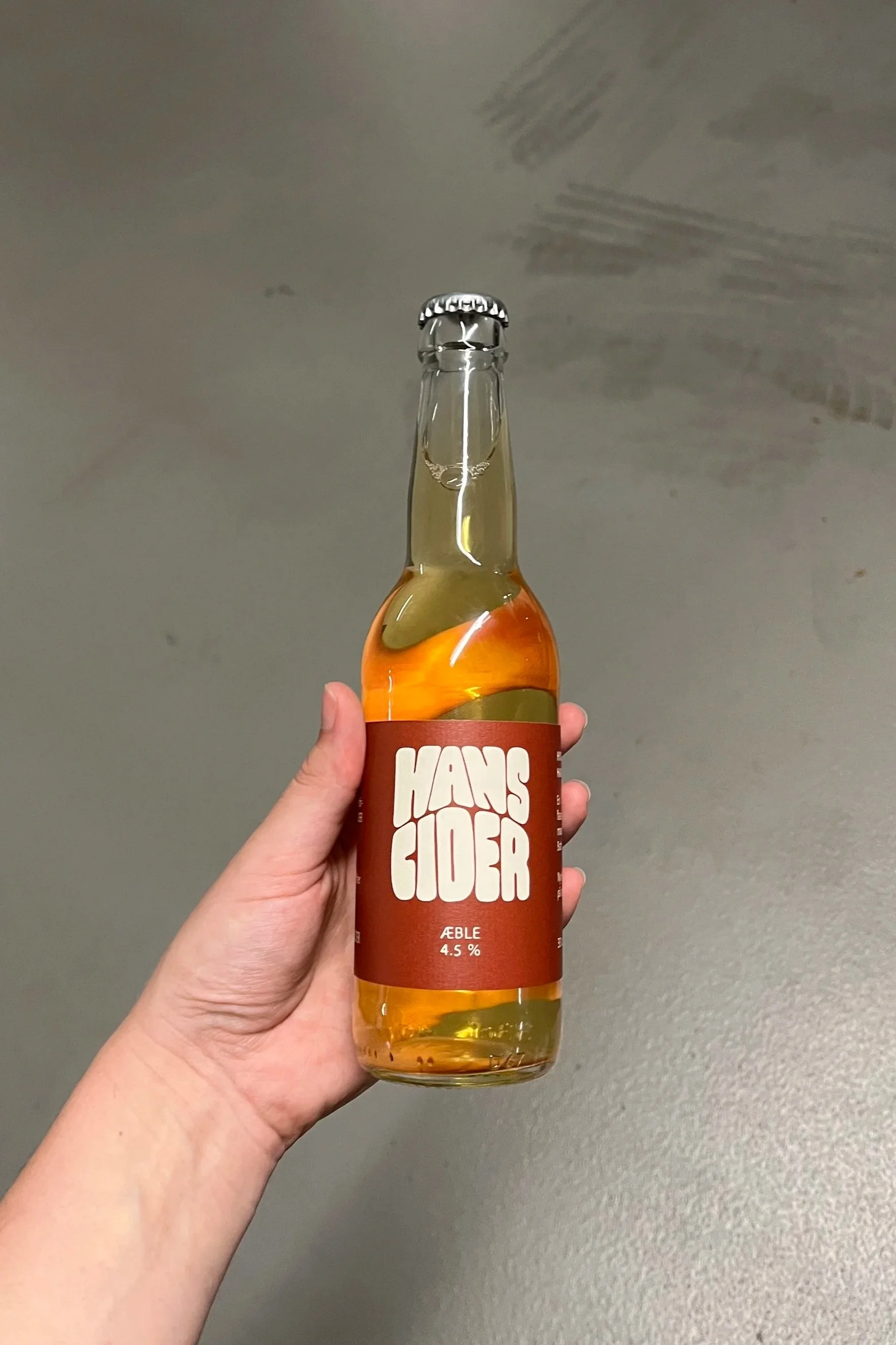 En person holder en flaske æblecider med mærket "Hans Johan" og 4,5 % alkohol. Flasken er gennemsigtig med gul væske og en orange mærkat.