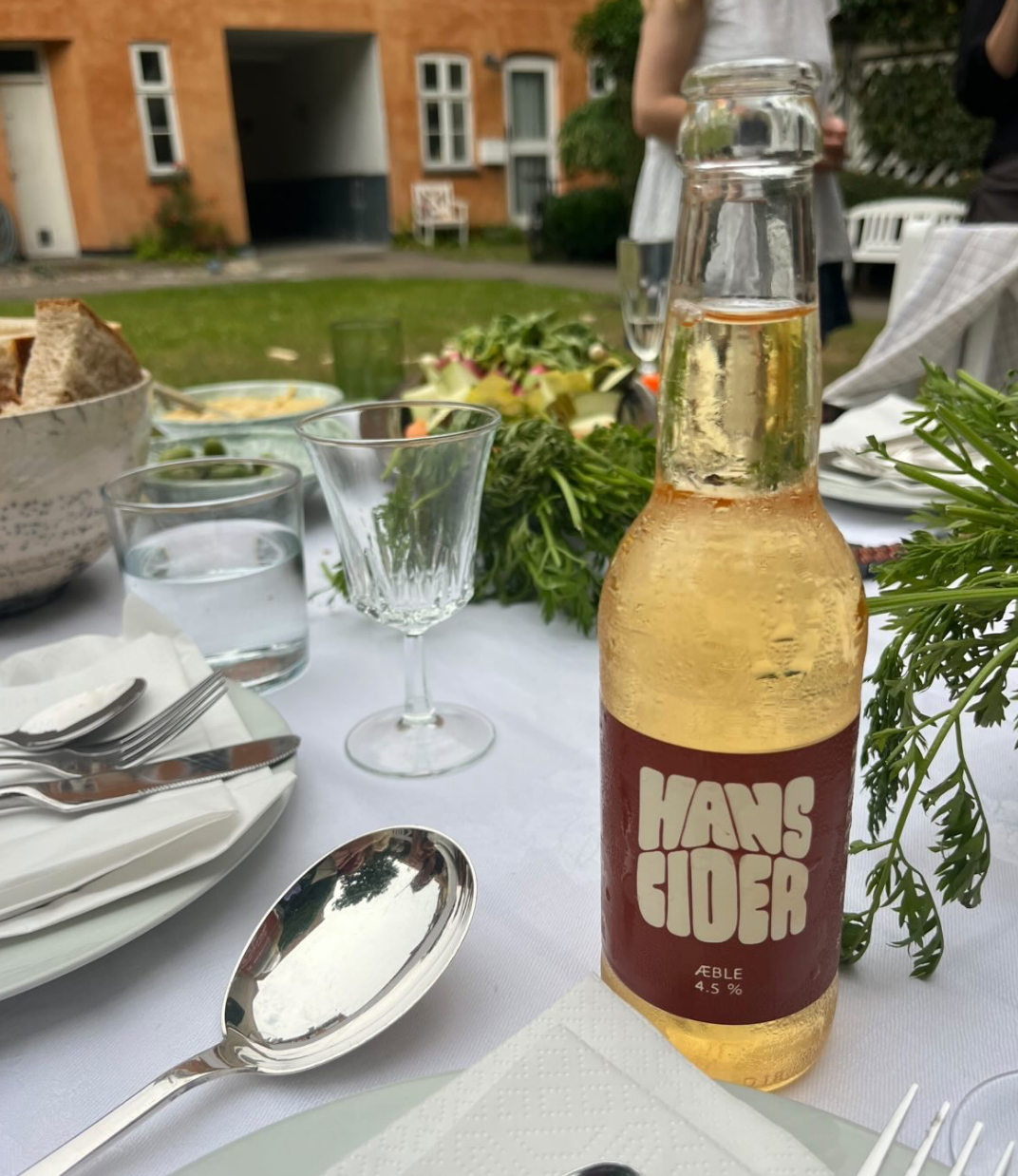En borddækket havefest med en ølflaske, glas, ske, tallerkener, brød, salat, og nydelig udeliv i baggrunden.