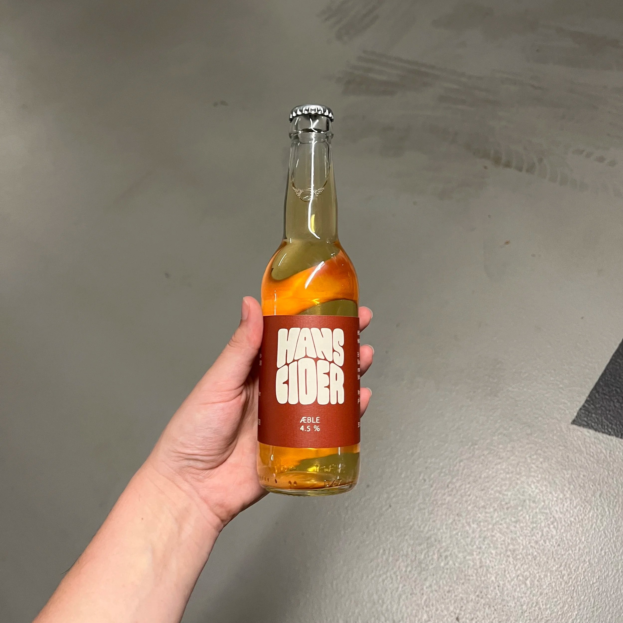 Hånd holder en glasflaske med æblecider, etiketten siger 'HANS HAS FLUER' og angiver 4,5% alkohol. Flaske er tomme, men væske er synlig i bunden.