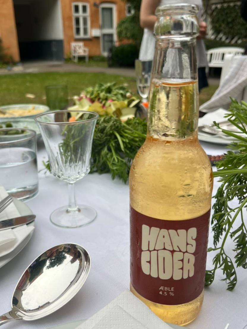 En glasflaske med Hans Jøder æble cider, 4,5 % alkohol, står på et bord dækket med en hvid dug under en udeservering.
